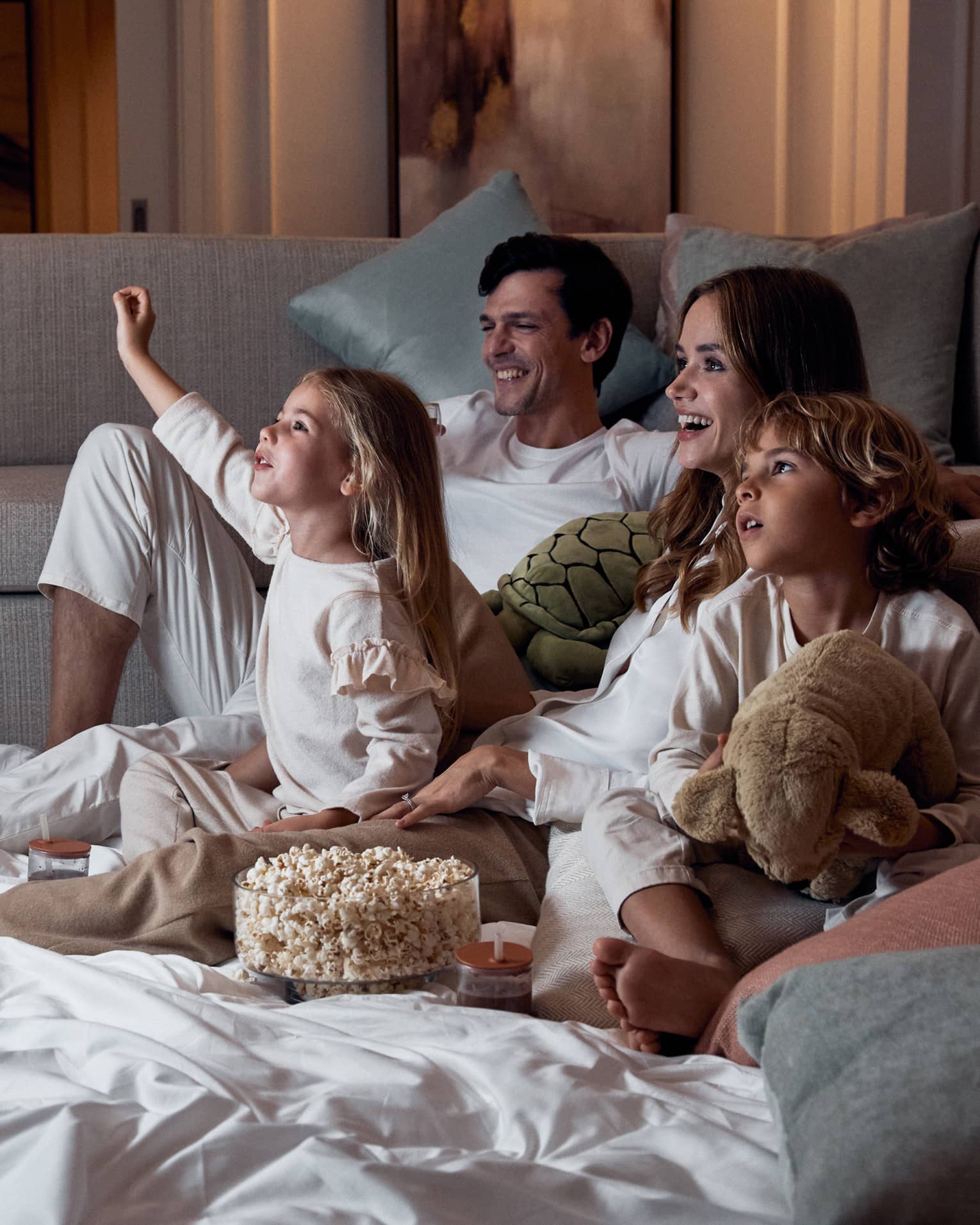 A family relaxing on blankets beside a sofa; the children hold stuffed toys and there's a bowl of popcorn between them.