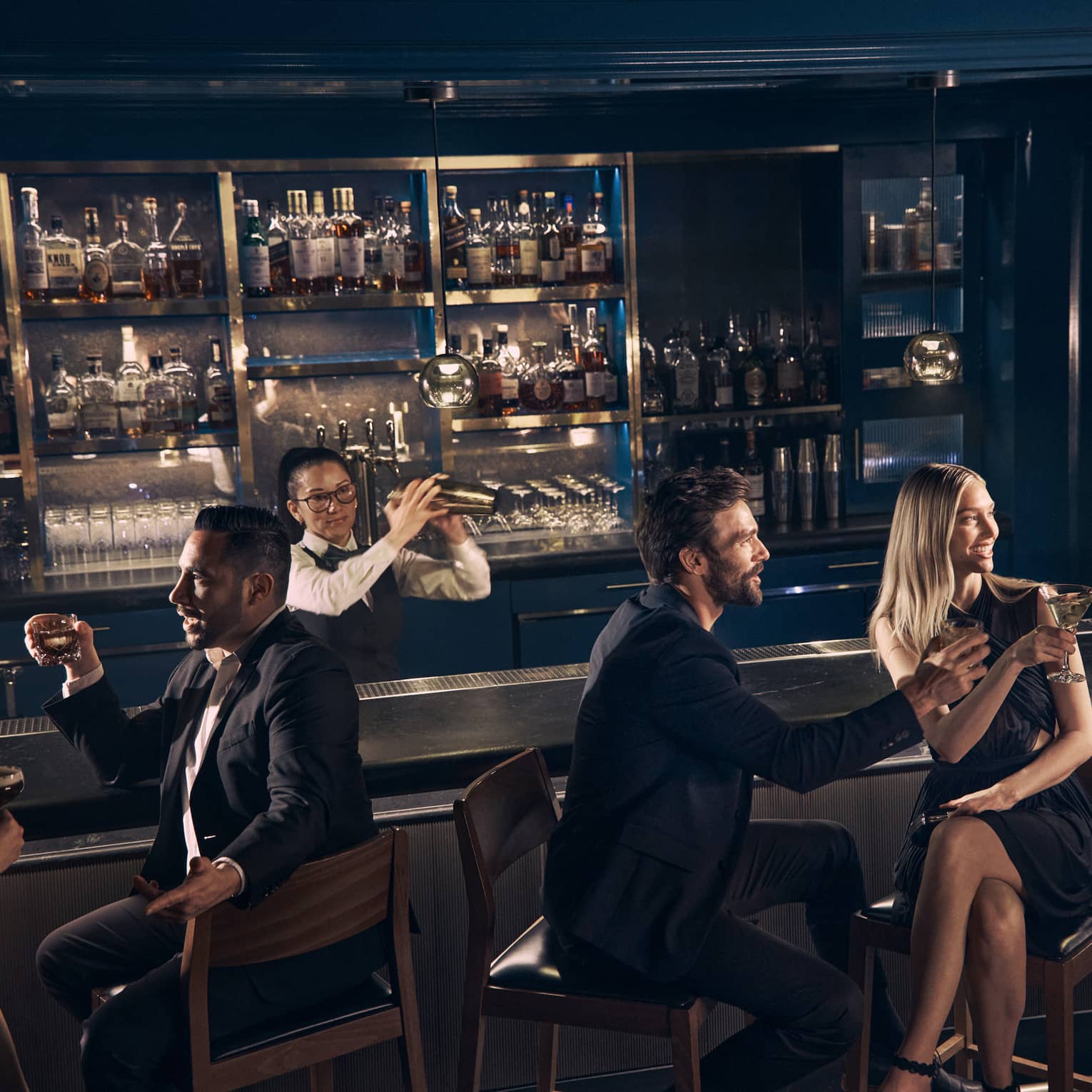 Guests chat over drinks at the bar at Prosperous Penny as a bartender shakes a cocktail shaker behind the bar.
