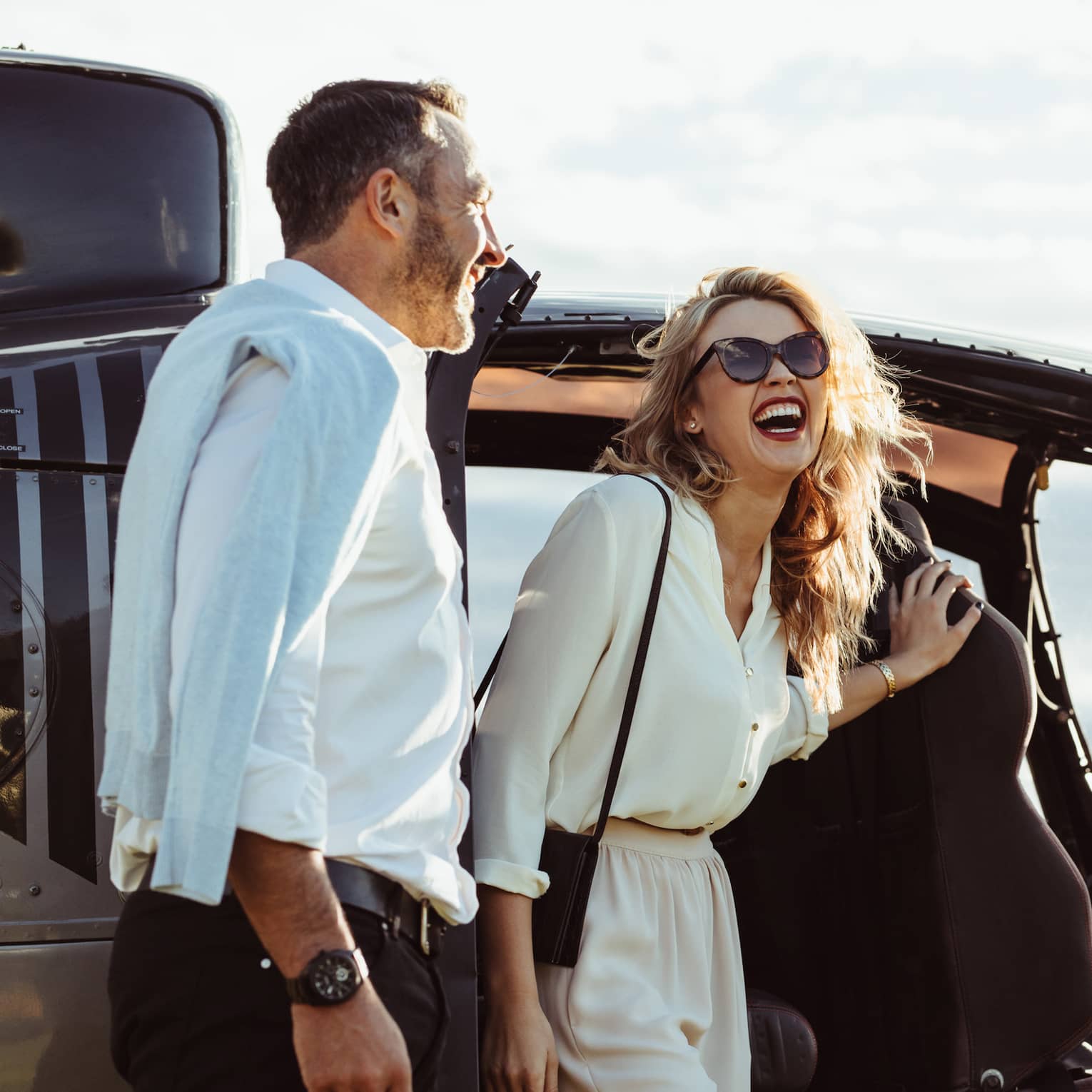 A man and woman laughing while getting out of a helicopter with the pilot smiling nearby.