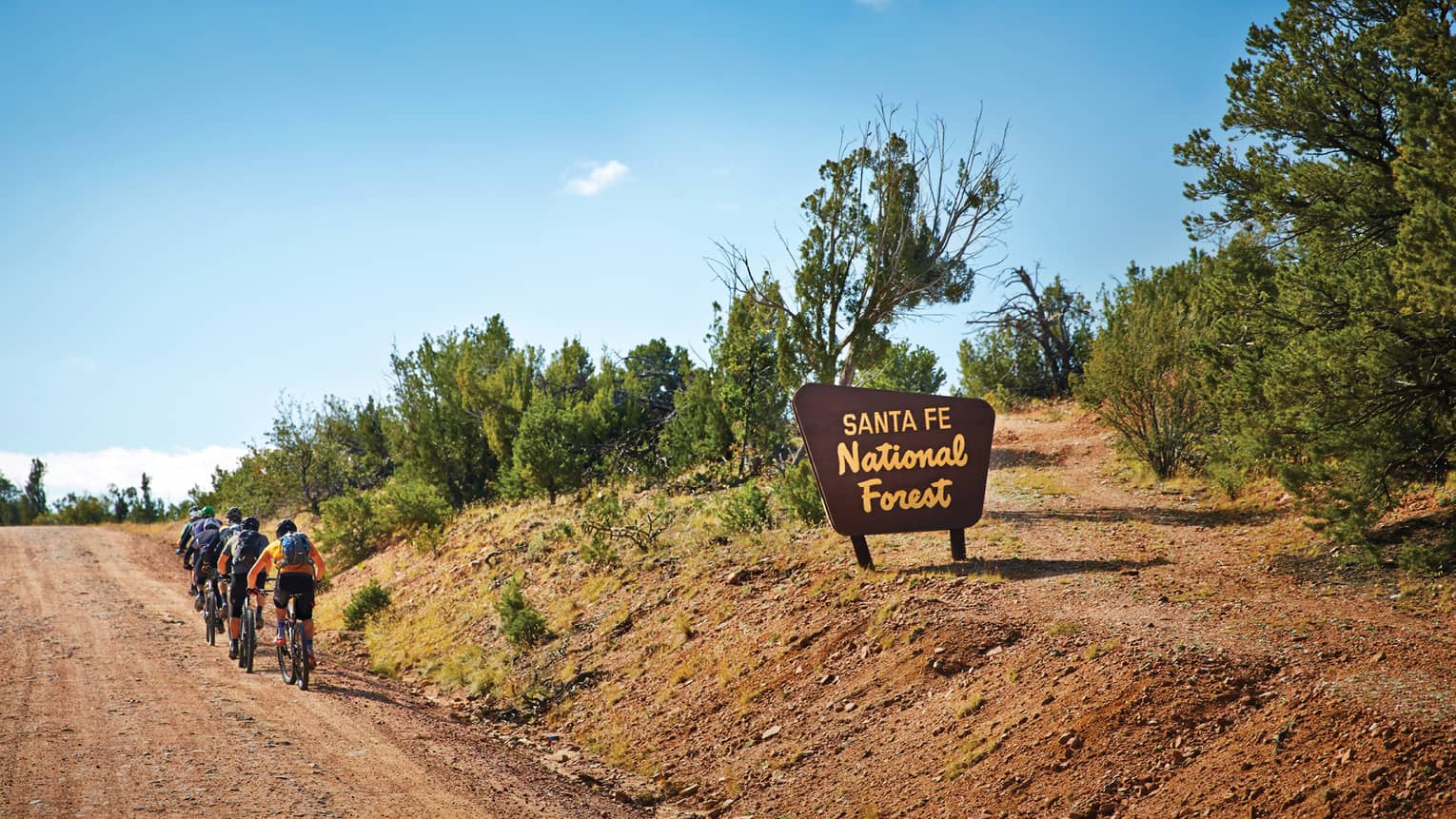 Group of cyclists ride up dirt path in a row past Santa Fe National Forest sign 