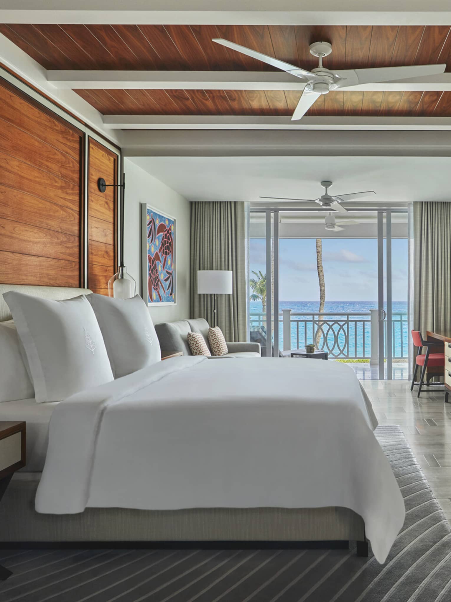 Ocean-view luxury resort bedroom with wooden accents, king bed, seating area, desk, wall-mounted TV and balcony