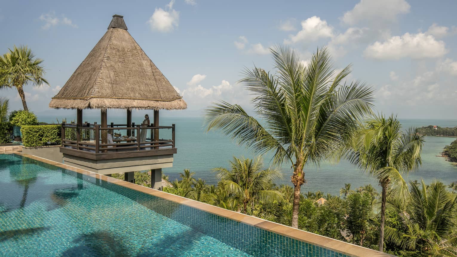 Thatched-roof pavilion at edge of outdoor swimming pool overlooking tropical trees, mountain, ocean