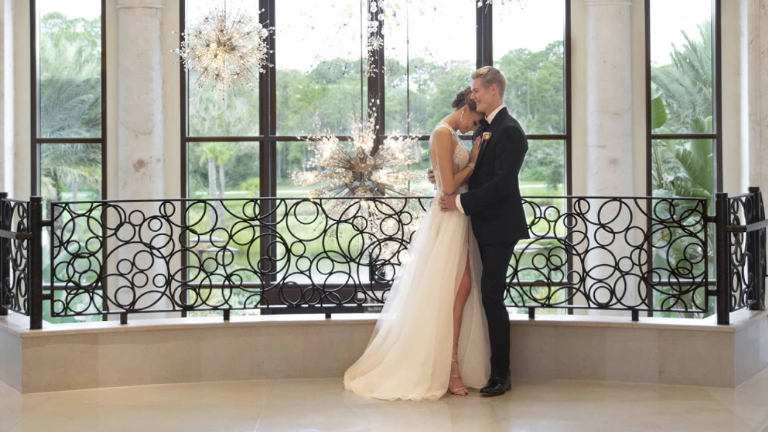 Bride and groom embrace by balcony overlooking white marble floors and wall, large window and modern chandeliers