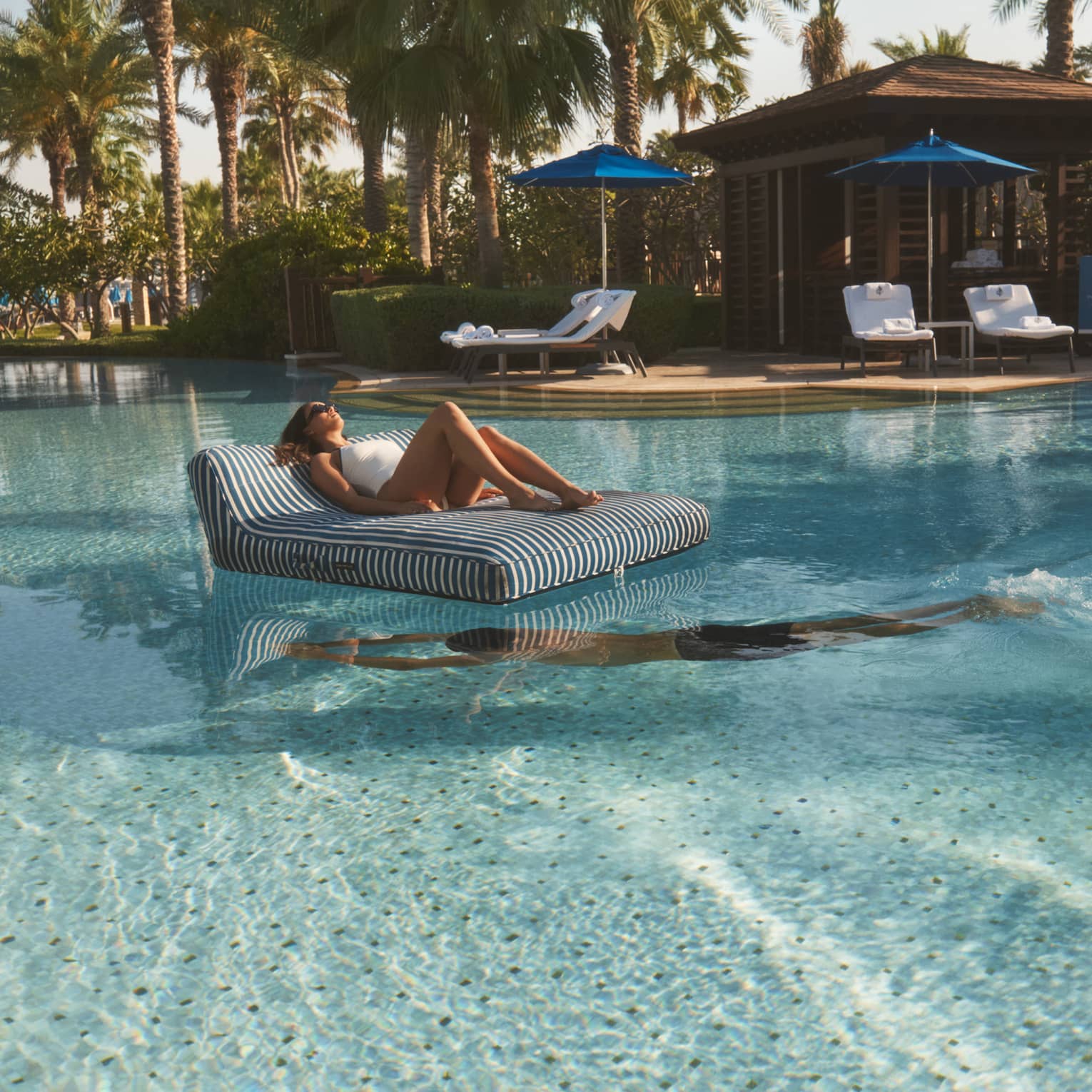 Person lounges on a black-and-white striped floating chair in the middle of a pool as another person swim beneath