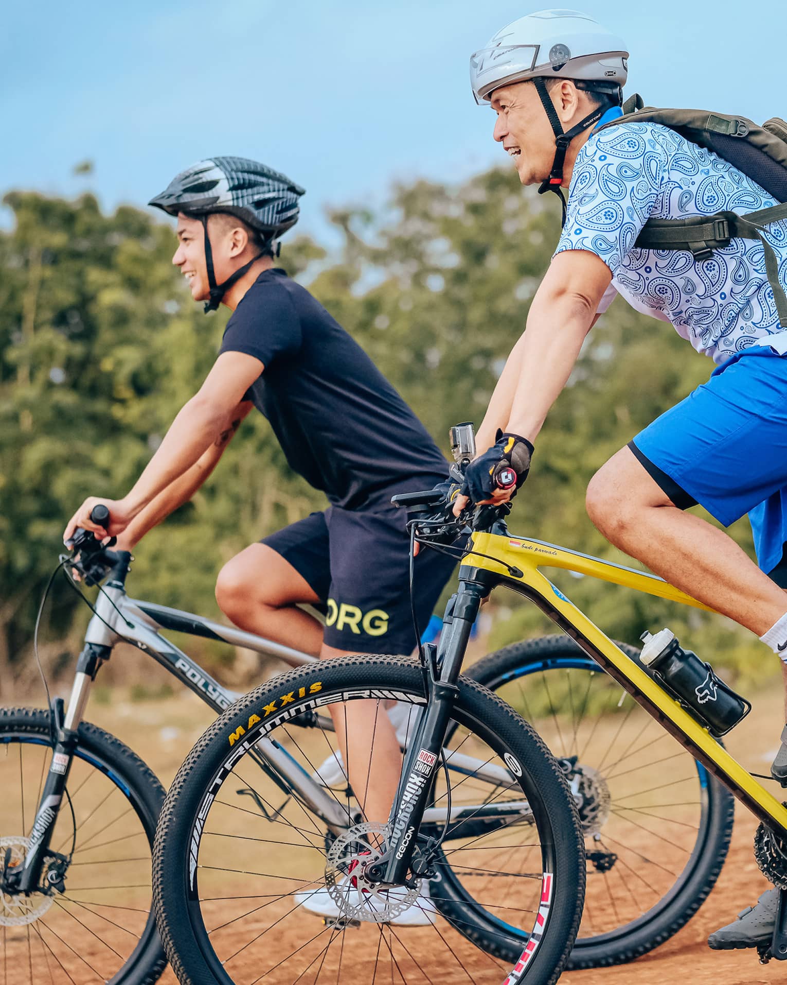 Side view of two smiling mountain bikers in helmets as they coast down a gentle slope on a dirt path alongside trees.