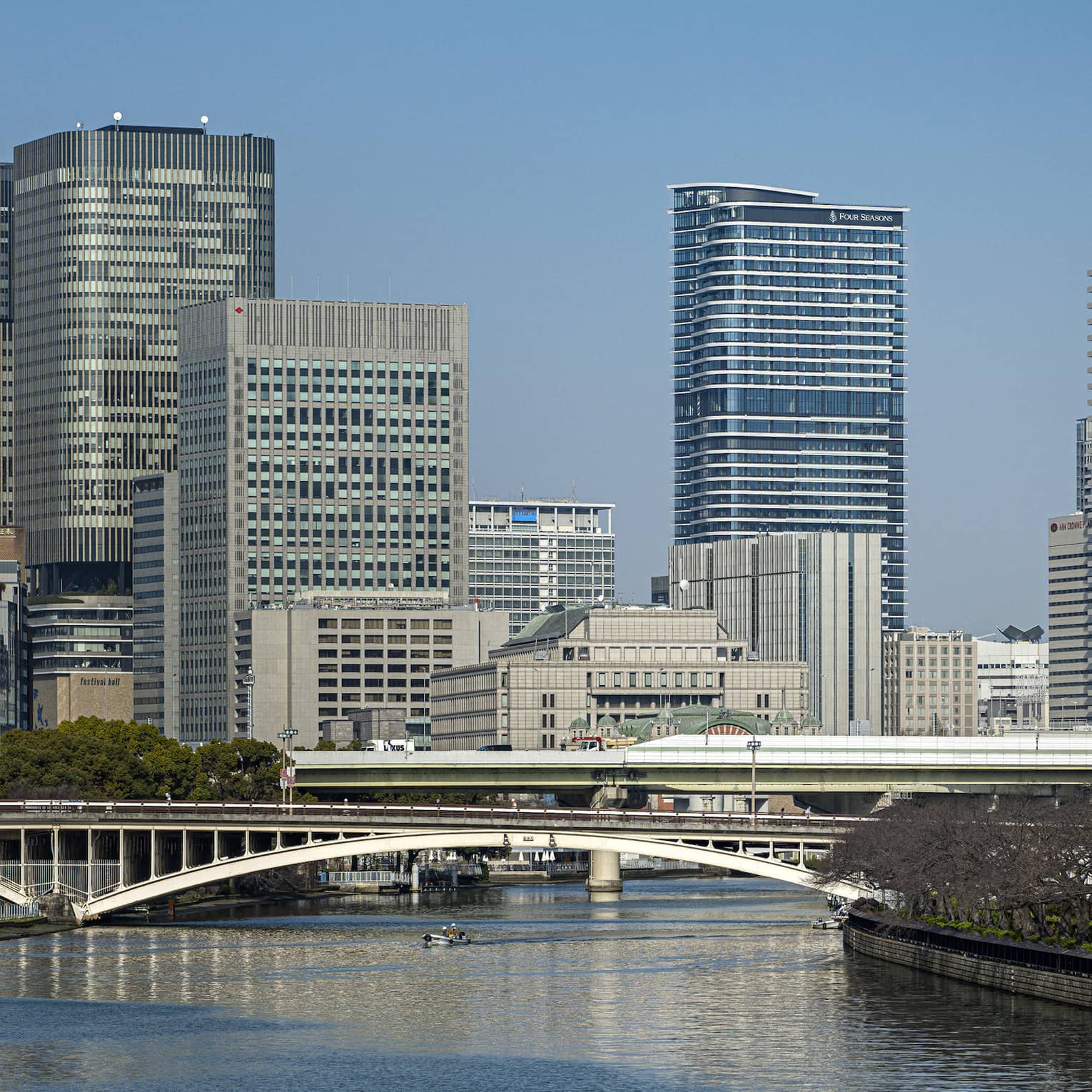 Under a clear blue sky, Osaka's modern high-rises loom above two bridges spanning a river lined with dormant cherry blossoms.