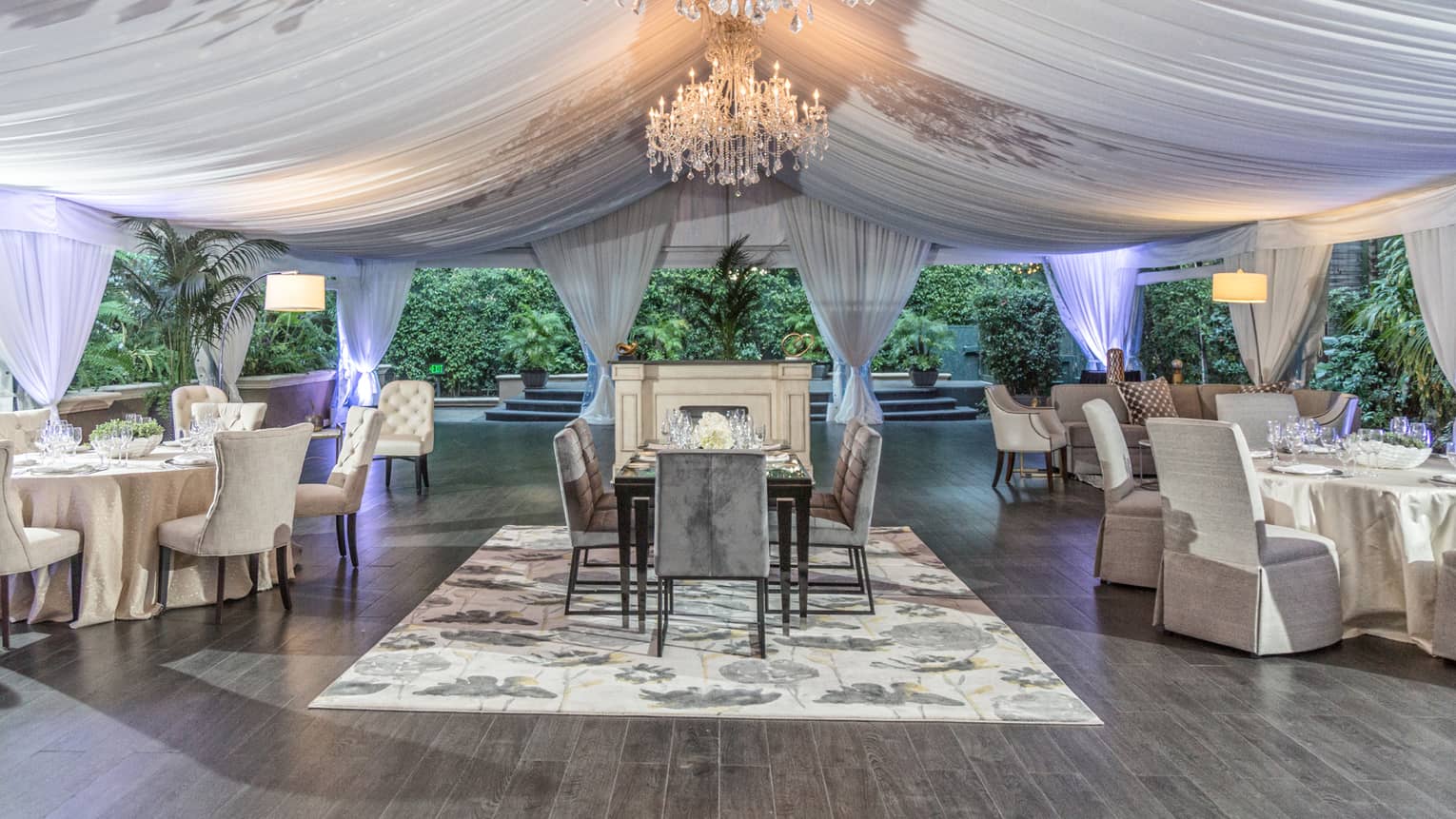 Chandeliers hang from a tent draped with cloth above tables with place-settings 