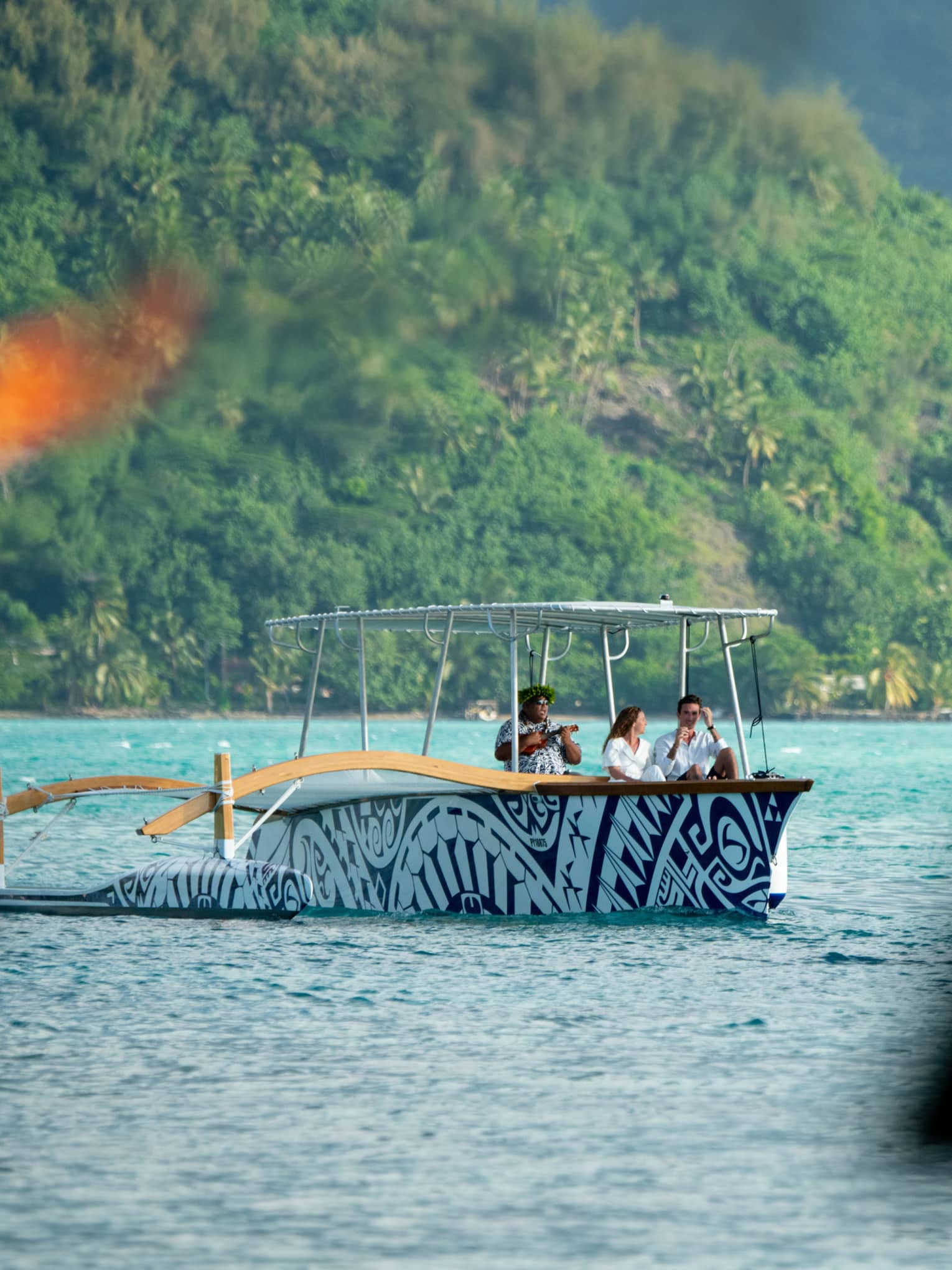A motorized outrider canoe carries a couple and a musician across the ocean; in the foreground torches flicker with fire.