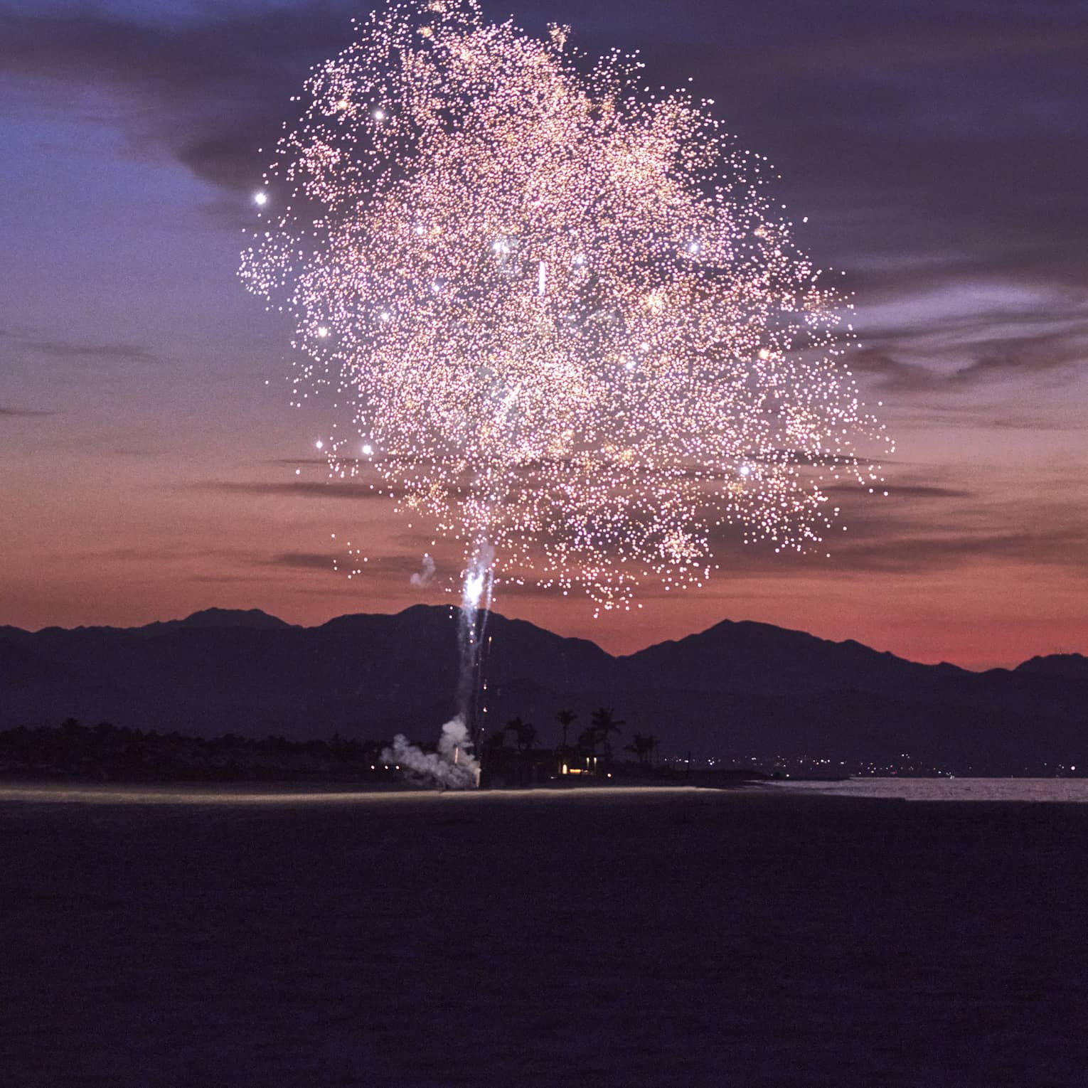 Pink fireworks exploding during sunset with a mountain behind them.