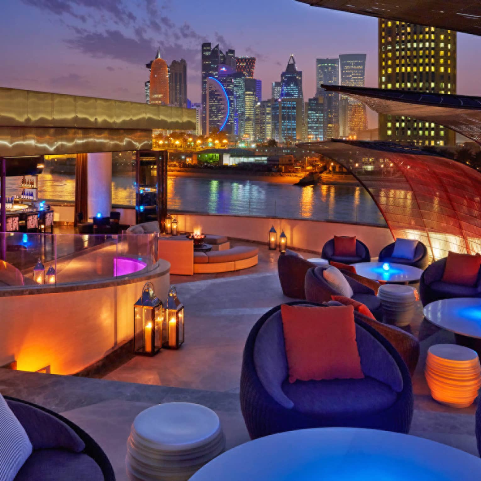 Rooftop bar furnished with round tables, plush chairs, lanterns, a curved bar area and a city skyline in the background