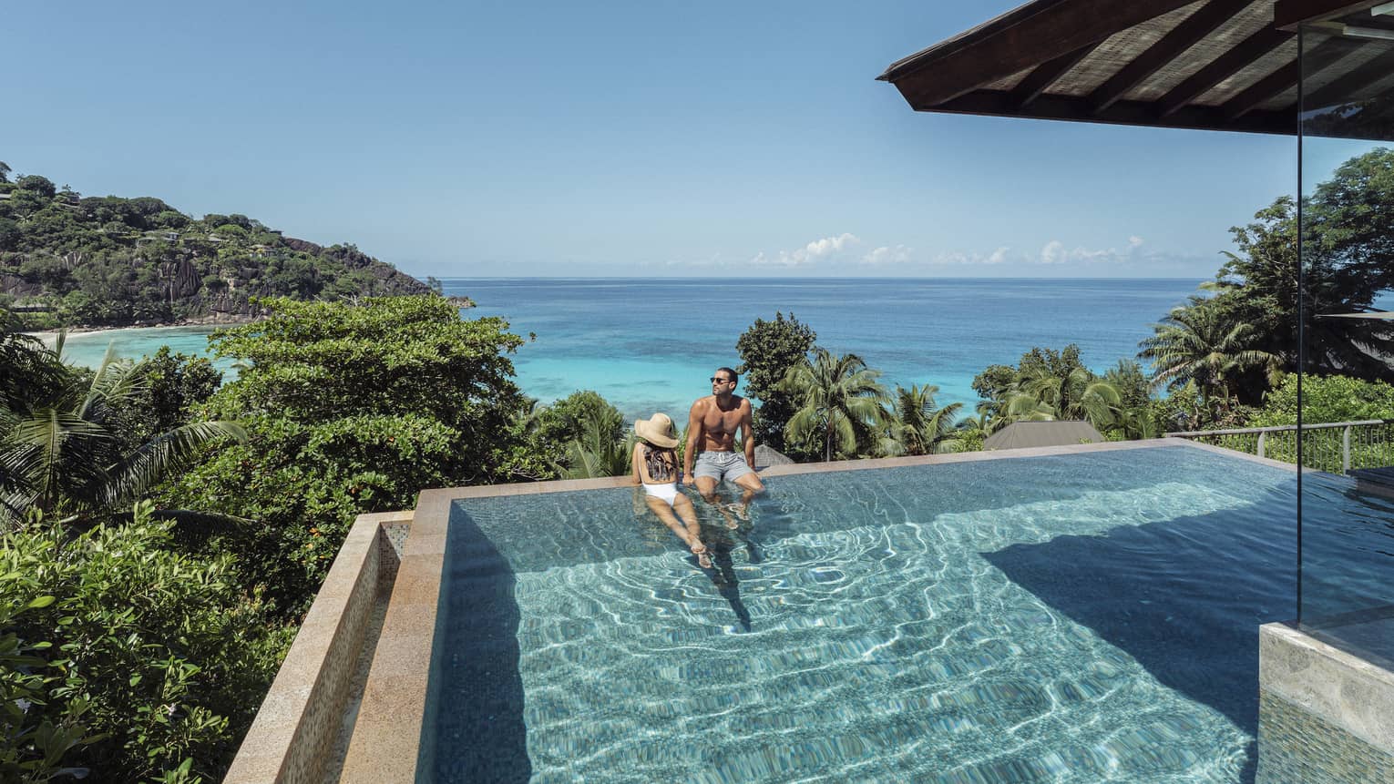 A couple lounges in a large infinity pool overlooking crystal blue waters