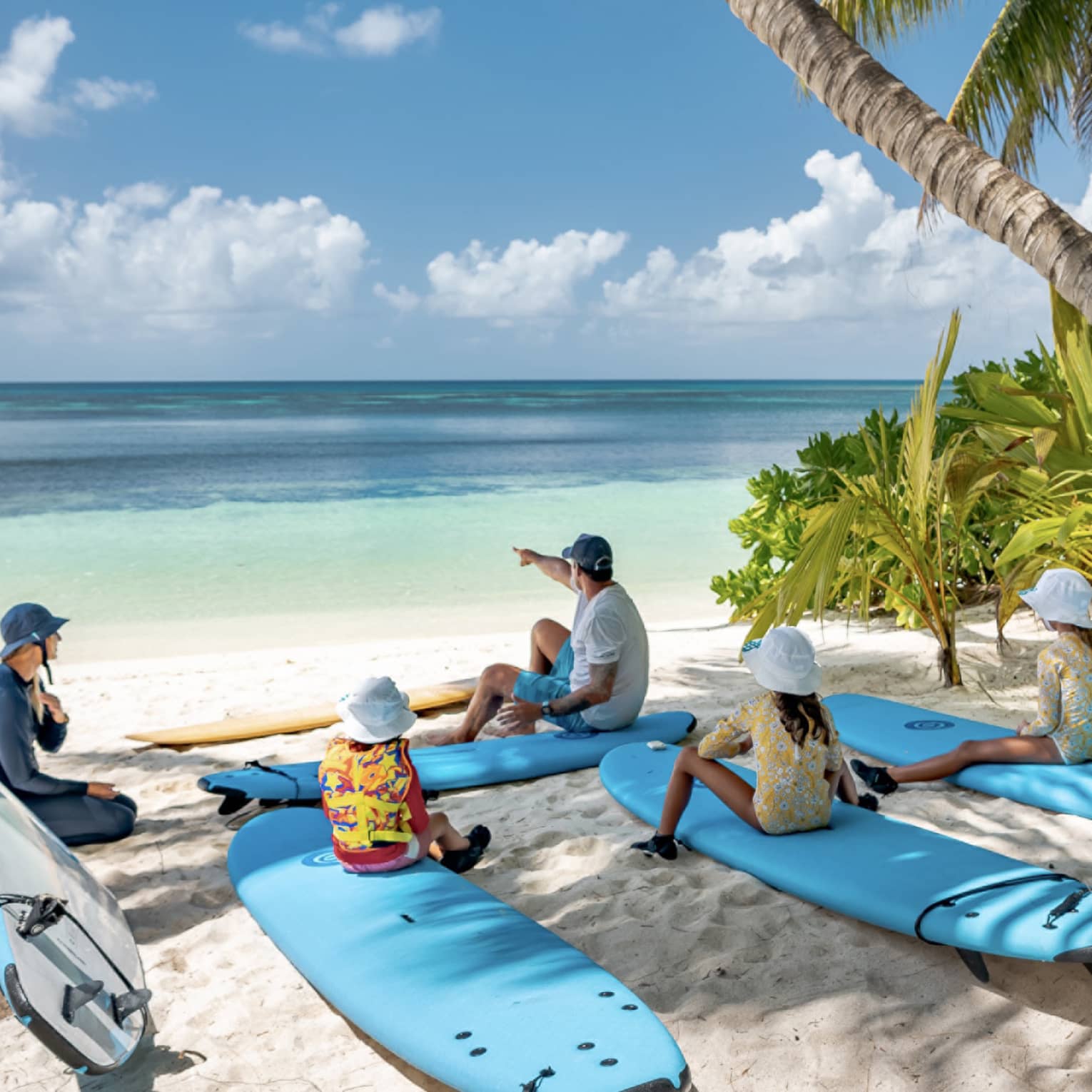 Children sit on surfboards under the shade of palm trees at the beach. One of their two instructors point at the clear water.