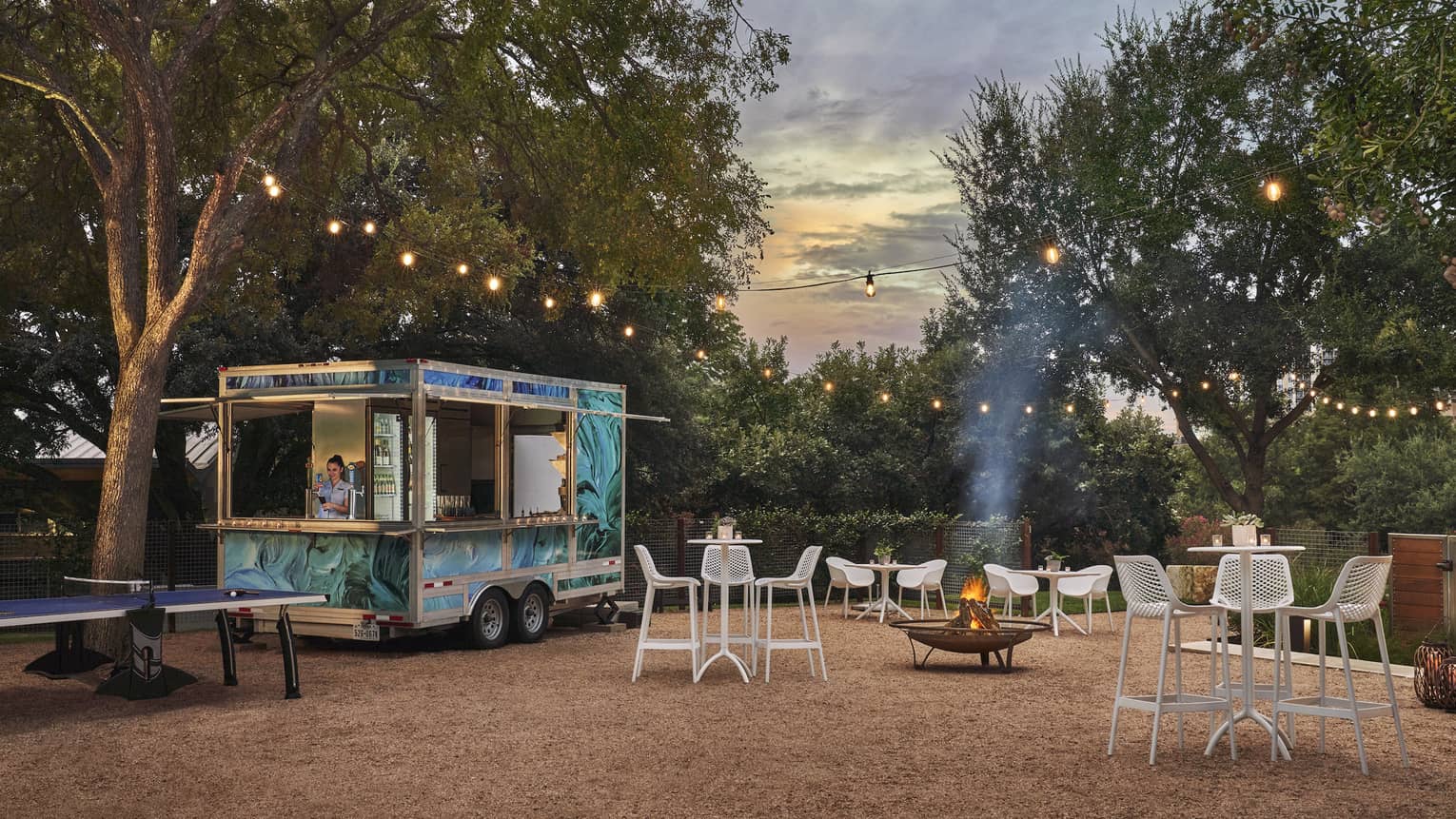 A food truck outsider surrounded by chairs, tables and trees.