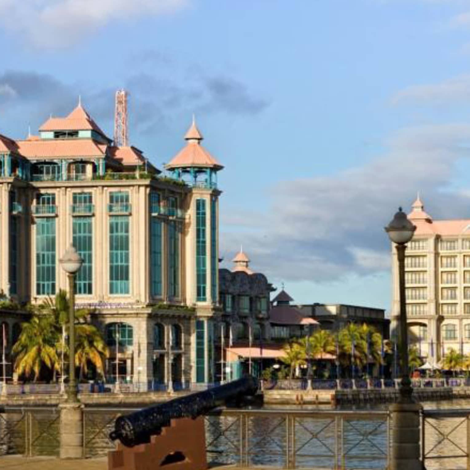 An old cannon in the foreground borders a channel, with ornate Colonial-style buildings across the channel on the shoreline.