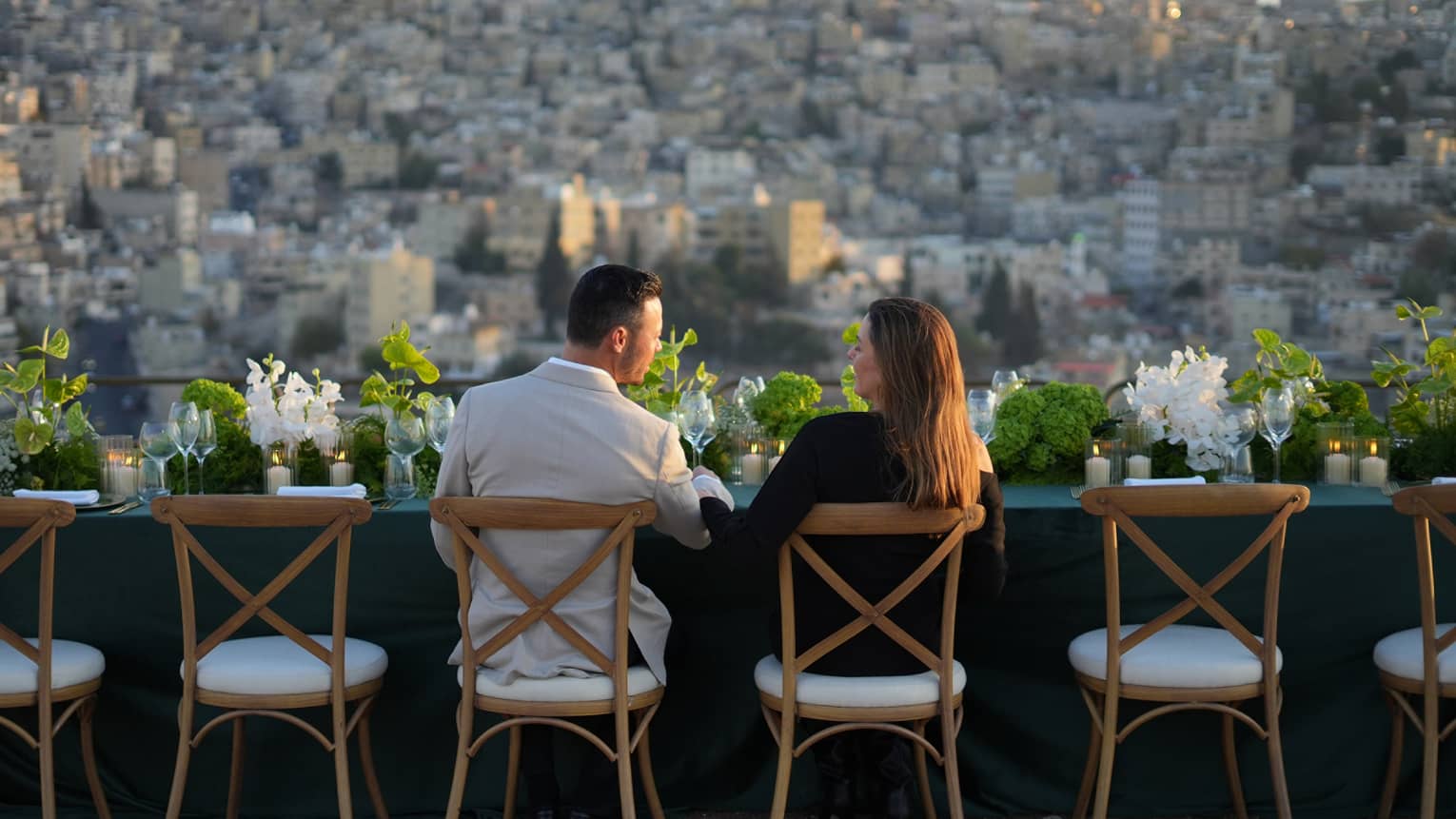 A couple sits at an outdoor table topped with floral arrangements while looking out at the city below