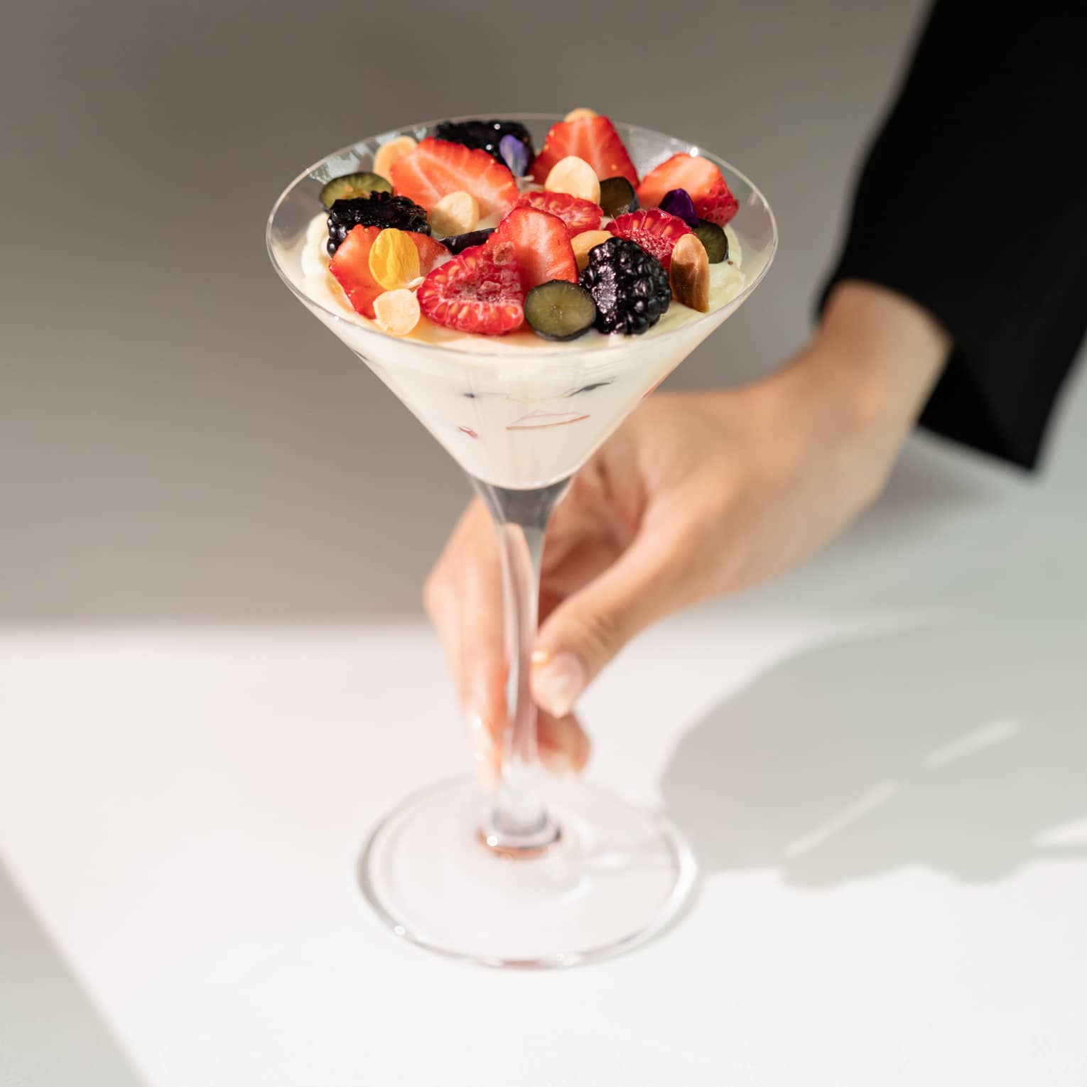 A hand delicately holds a sunlit martini glass filled with yogurt topped with summer fruits, chopped nuts, and flower petals.