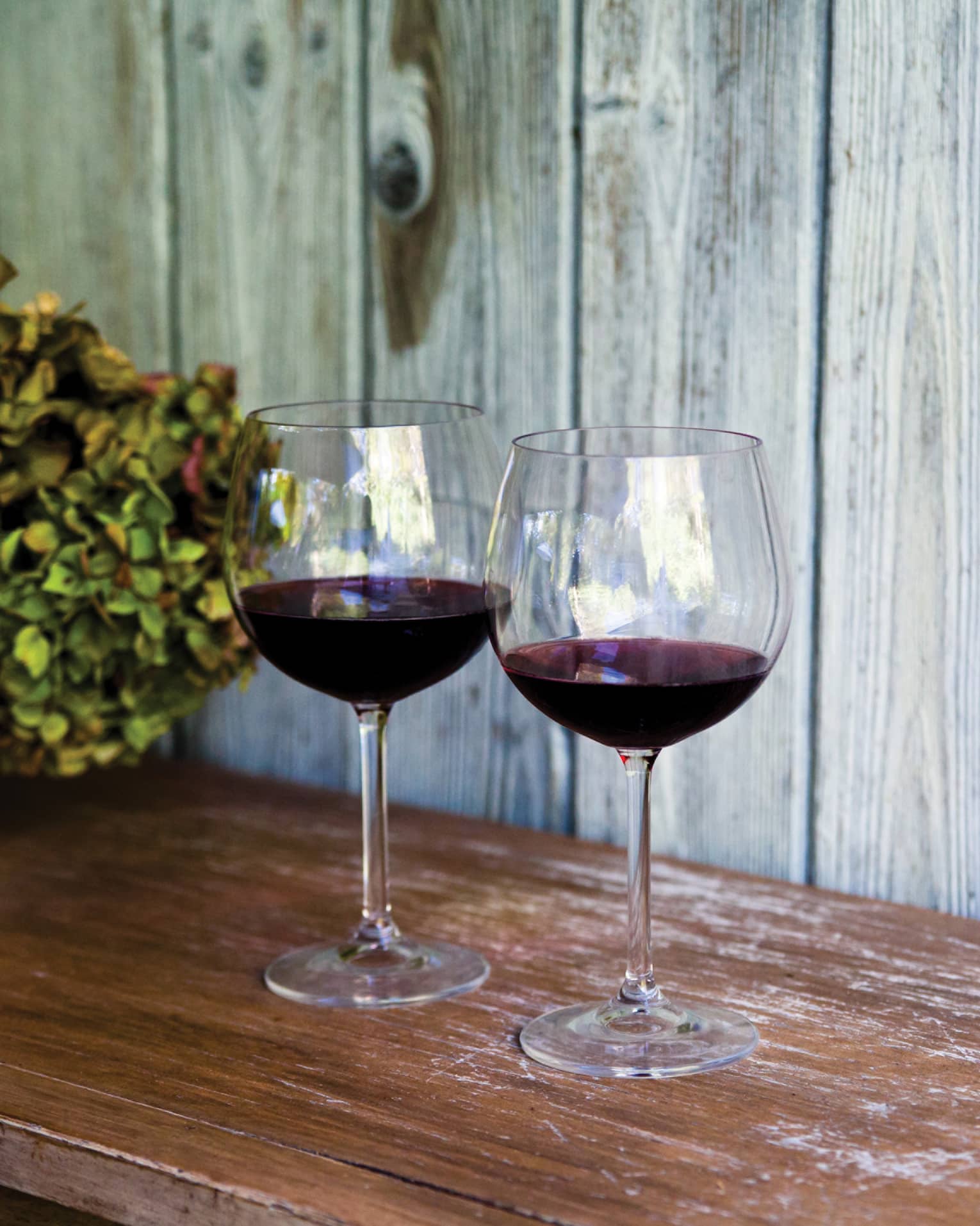 Two glasses of red wine and sit on wooden table, with white vase filled with hydrangeas 