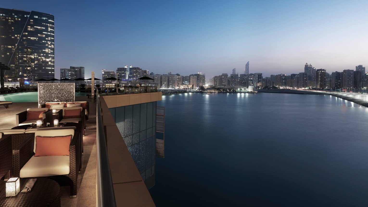 Hotel rooftop patio overlooking Arabian Gulf at night with wicker chairs with cushions, swimming pool