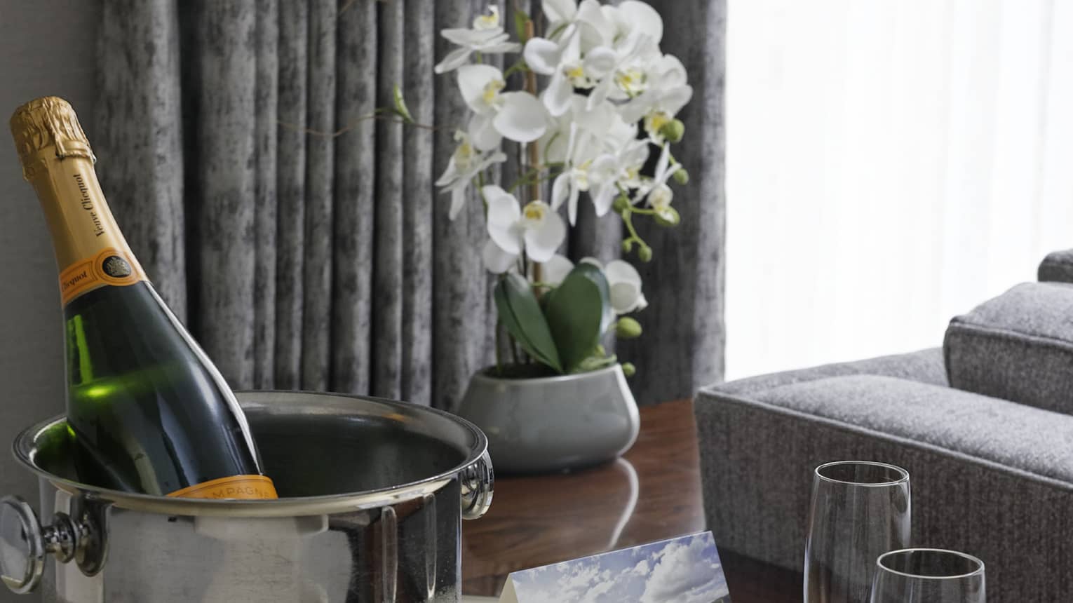 Ice bucket of Champagne and two flutes on round table, grey couch and window with grey curtains in the background