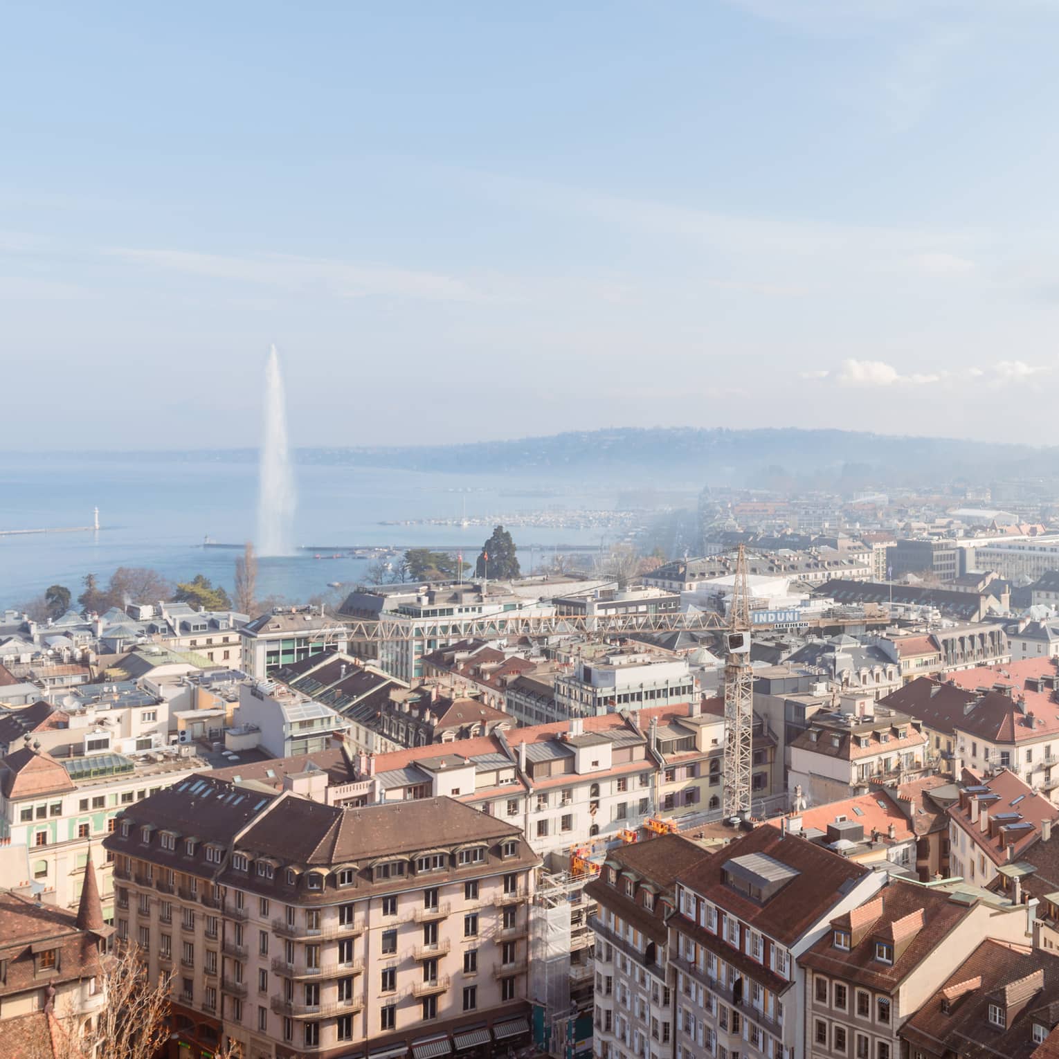 A cityscape with rooftops, buildings and streets, with a large fountain shooting water into the air over a lake in the background under a clear blue sky