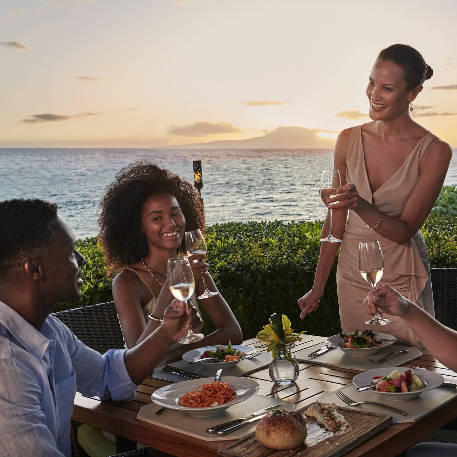 Two couples dine outside at Ferraro’s Bar E Ristorante, overlooking Wailea Beach. The sun is setting as they raise a toast. 