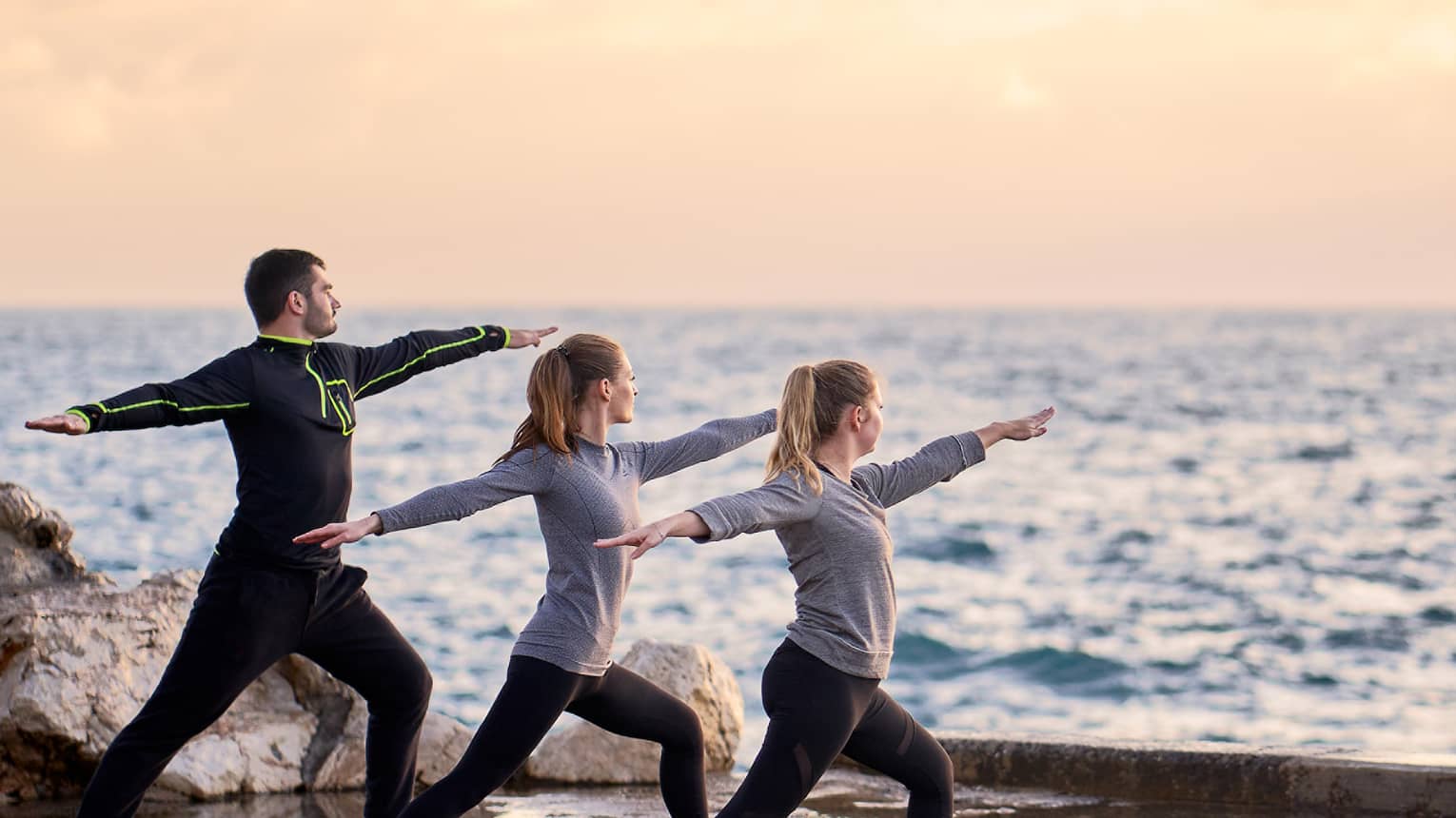 Three people in warrior yoga pose and coach standing at sea's edge at sunrise
