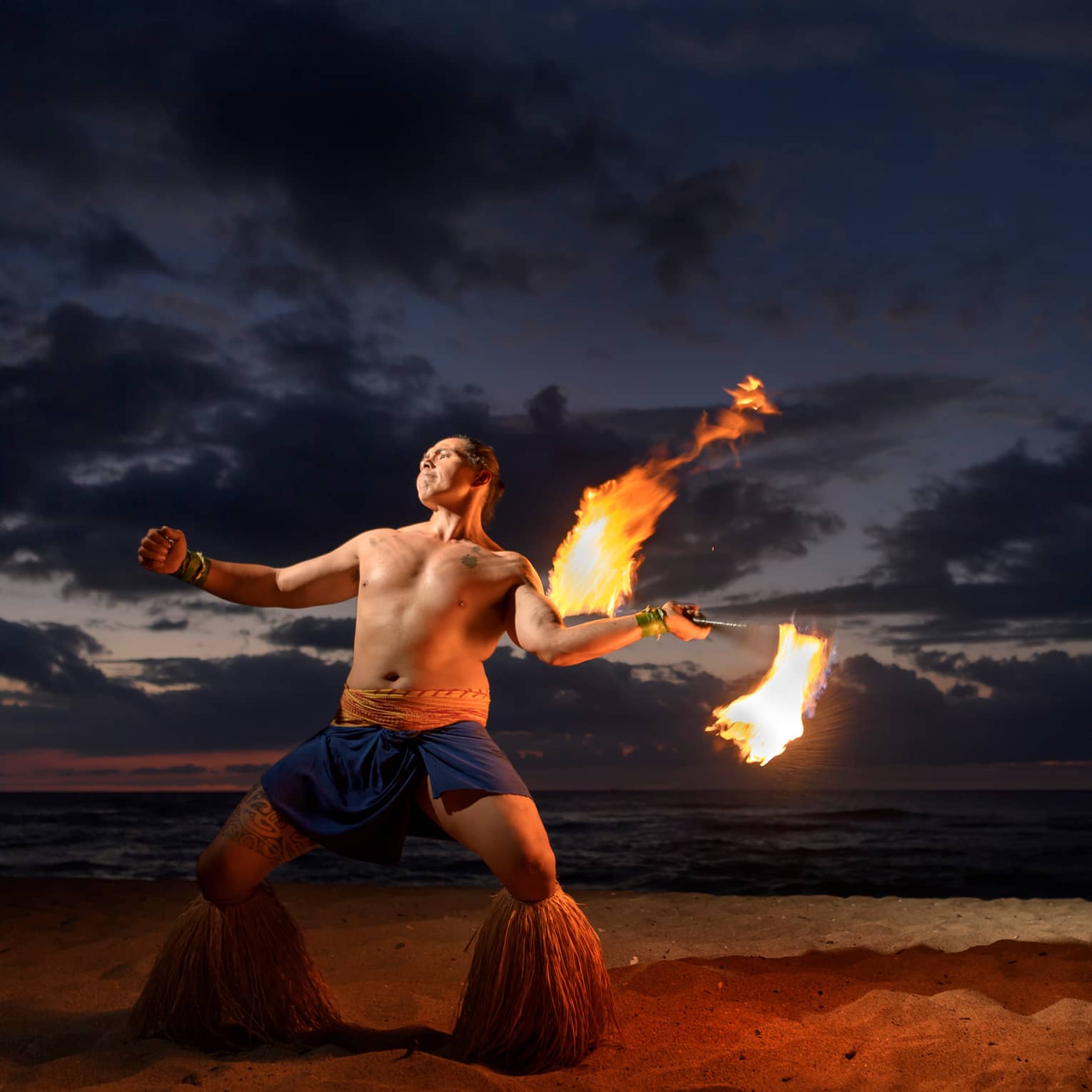 Man wearing traditional Polynesian outfit dances with torch on beach at night