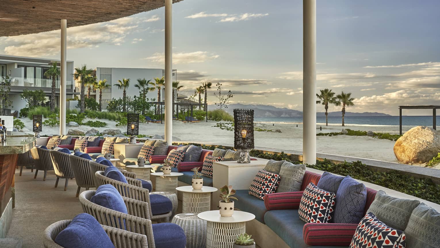 Out door lounge area at Casa de Brasa has built in couches with purple and pink cushions, opposite of rattan egg-shaped chairs overlooking the beach and ocean with the hotel in the background