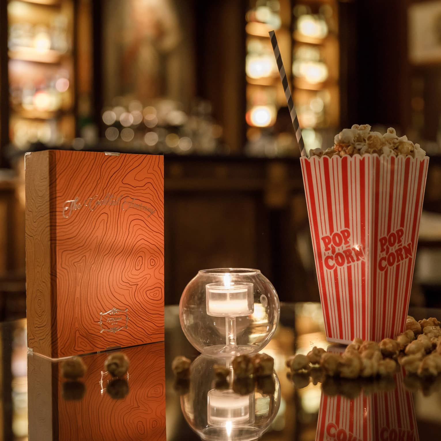 A red and gold box of chocolates, a candle in clear glass and a tall box of popcorn on a shiny table reflecting warm lights.