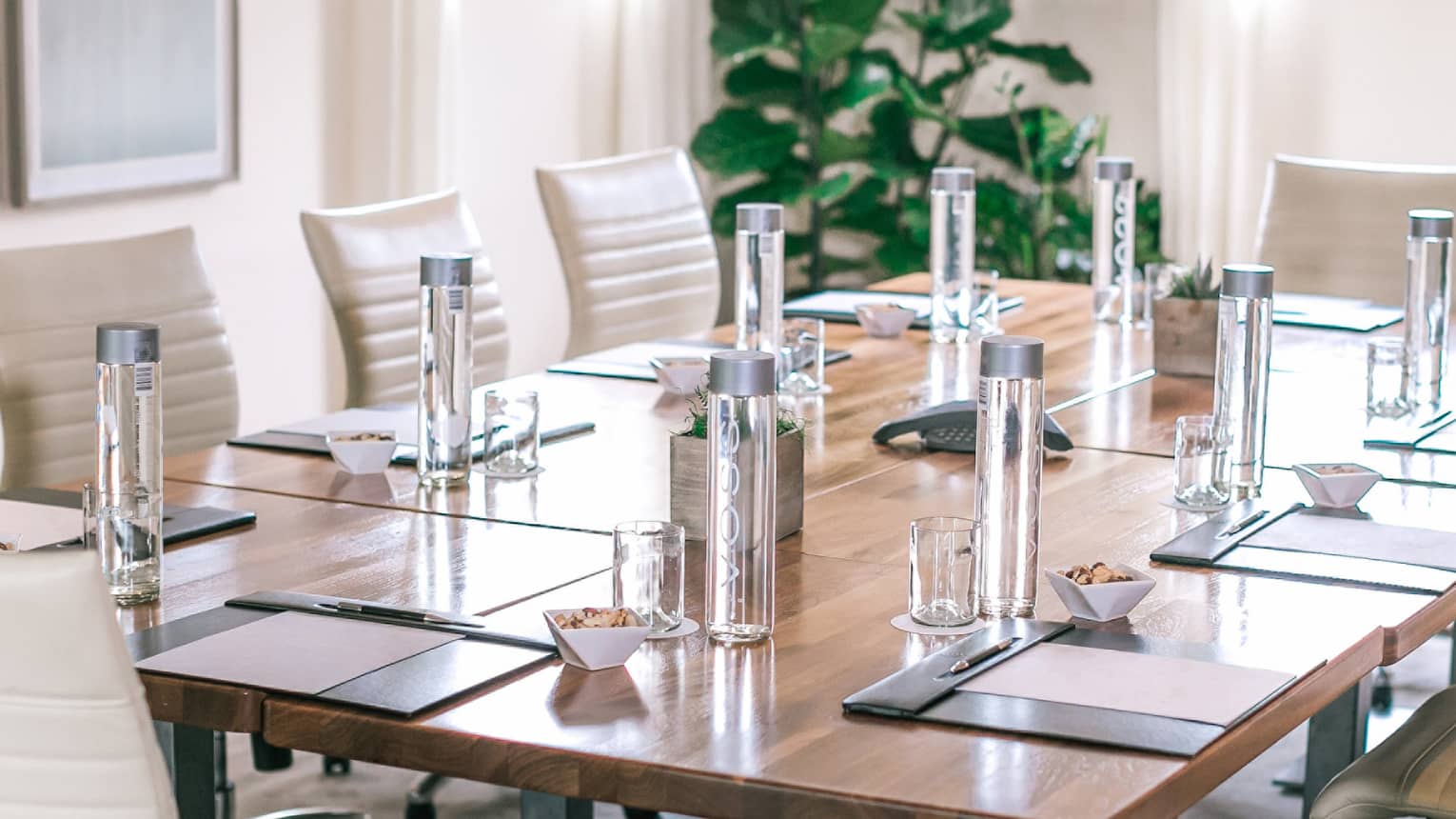 A rectangular boardroom table set with clear glass water bottles and black folders in a naturally-lit room