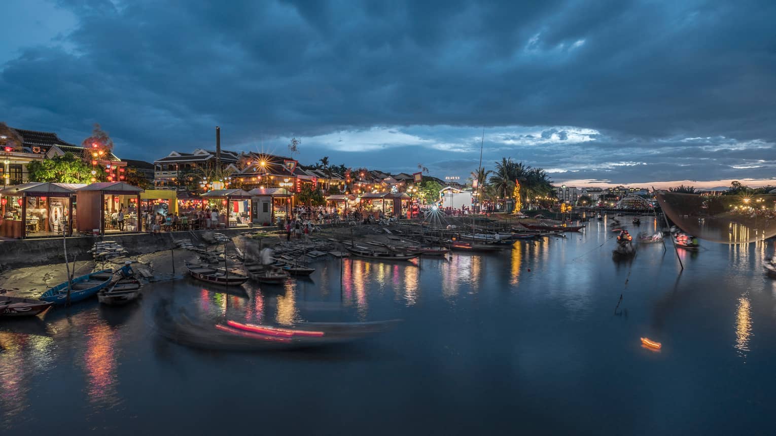 Evening view of a riverside market with colourful lights reflecting on the water and traditional wooden boats docked along the shore.