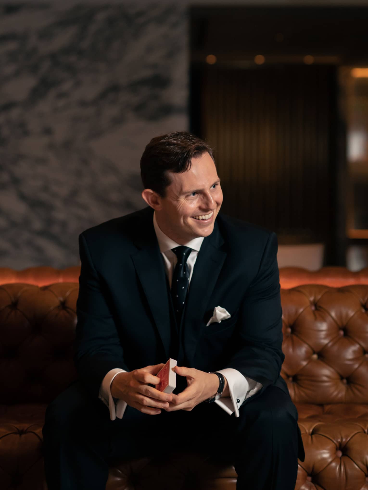 A man in a black suit sitting on a leather sofa holding playing cards.