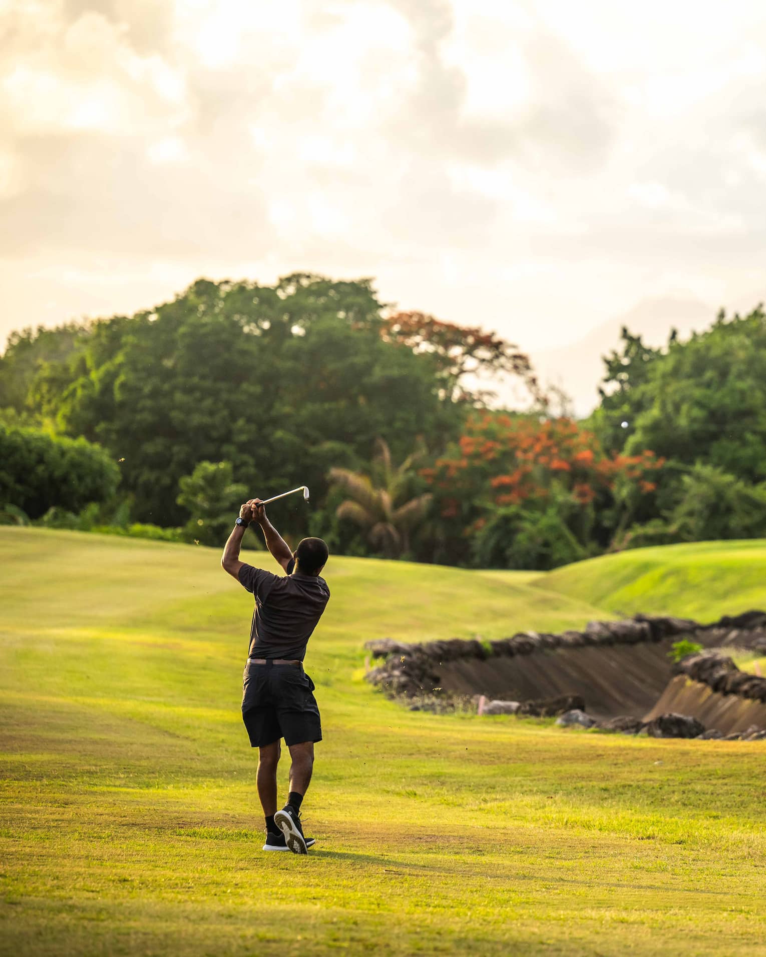 A golfer takes a swing on a bright green golf course surrounded by trees and lush vegetation, under a bright, partly cloudy sky