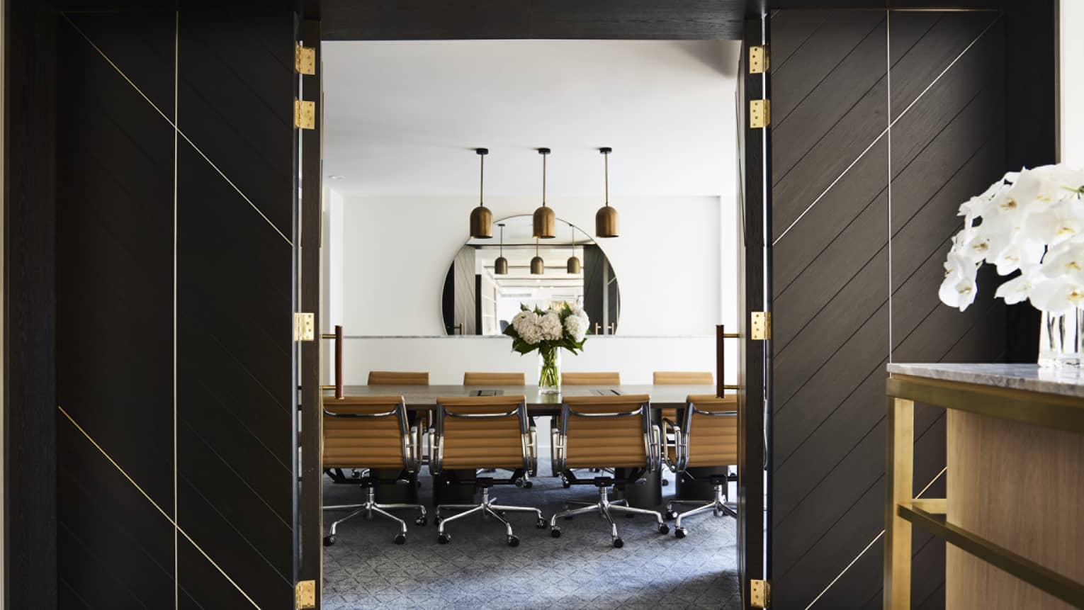 Tall open doors to private boardroom table, swivel chairs under three lights, mirror