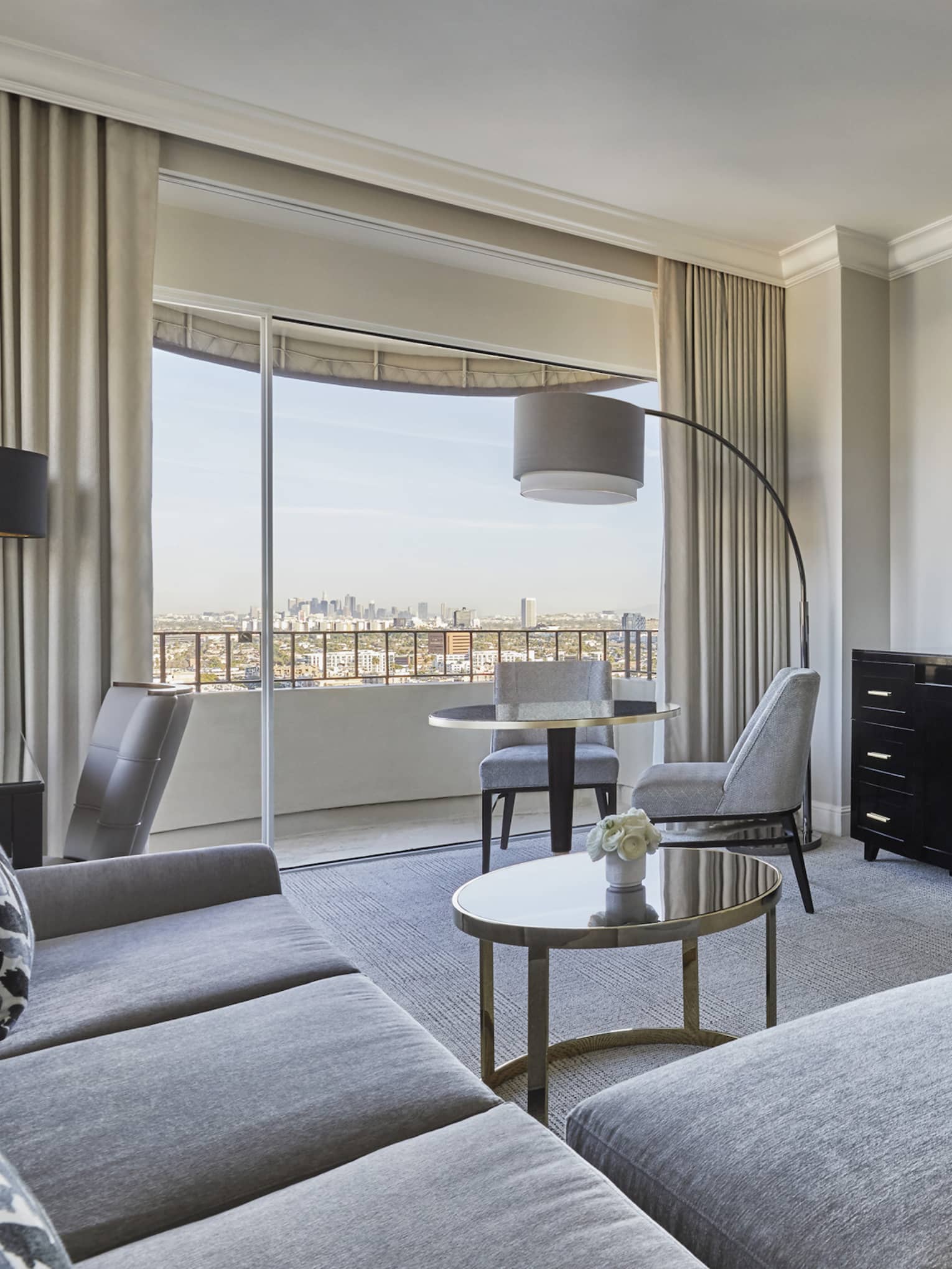 A hotel room with a grey couch, dresser, tv, lamp, small table, bed with white sheets, and large window.