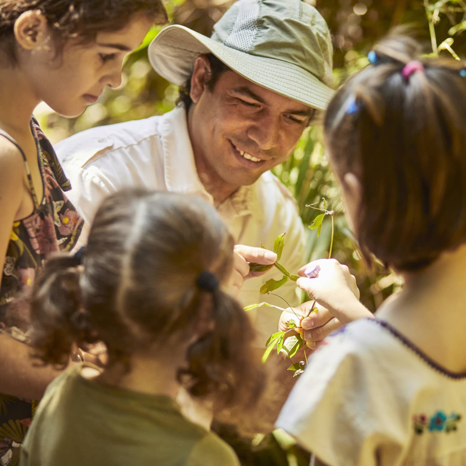 Three children gather around a guide wearing a sun hat as he shows them a plant. One child reaches to touch the leaves.