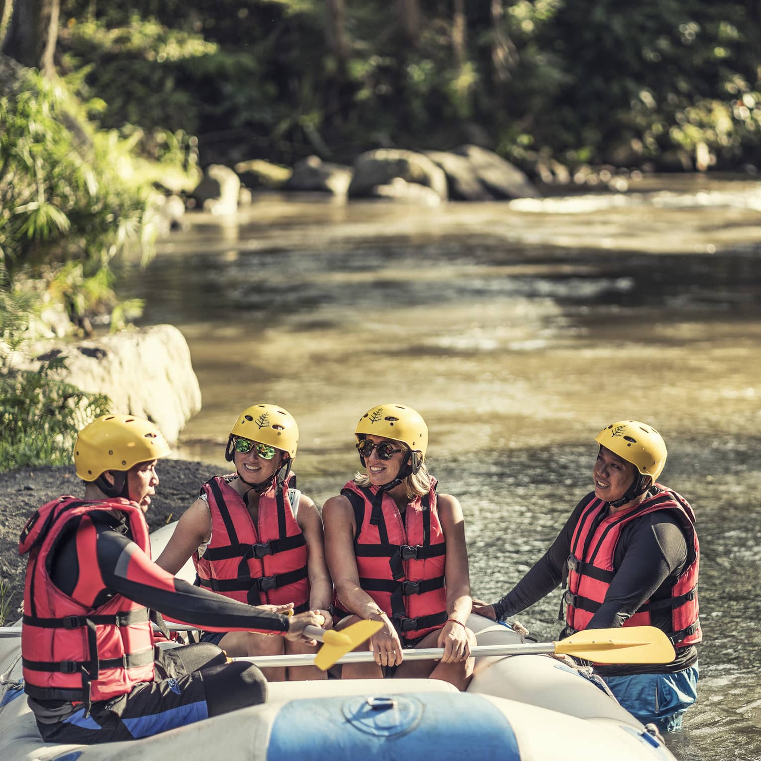 Four people wearing red life jackets, sunglasses and yellow helmets sit facing each other in a river raft