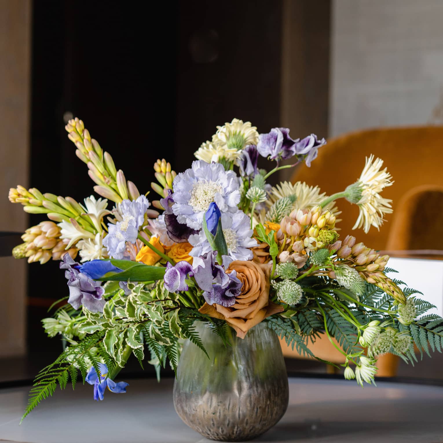 A lush, natural display of colourful flowers and greenery in a patterned glass vase, sitting on a marble table. 