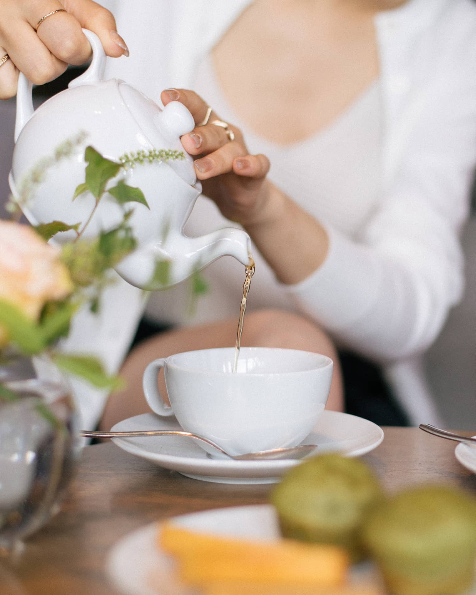 Two people sit at a table set for tea, with cups, saucers, a plate of small food items and a floral arrangement. One person is pouring tea from a teapot into one of the teacups.