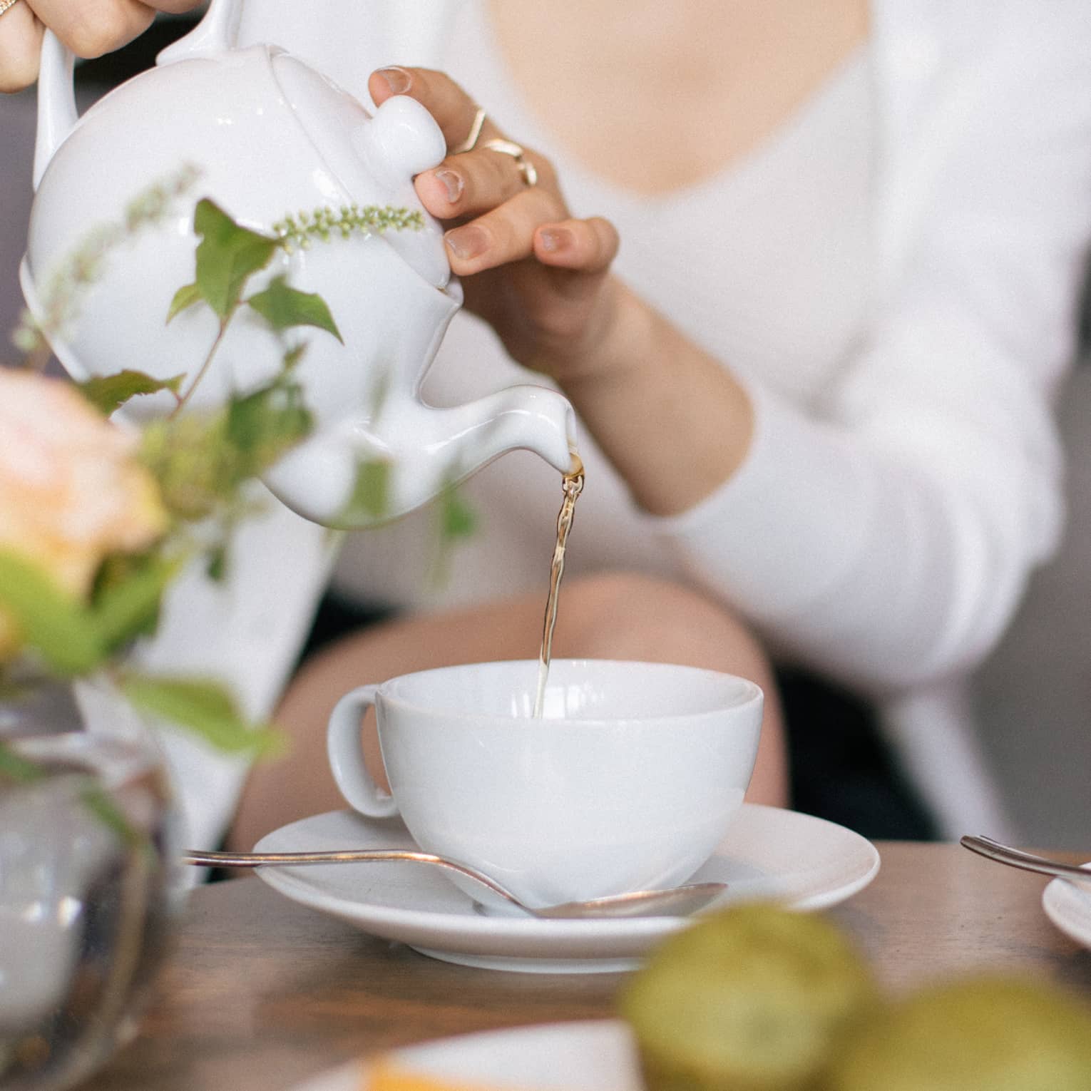 Two people sit at a table set for tea, with cups, saucers, a plate of small food items and a floral arrangement. One person is pouring tea from a teapot into one of the teacups.