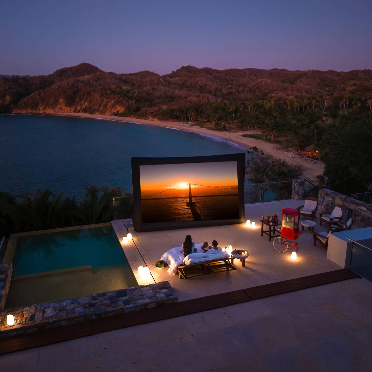 A couple watches a movie on a projector screen on a pool deck with a mountain view at sunset