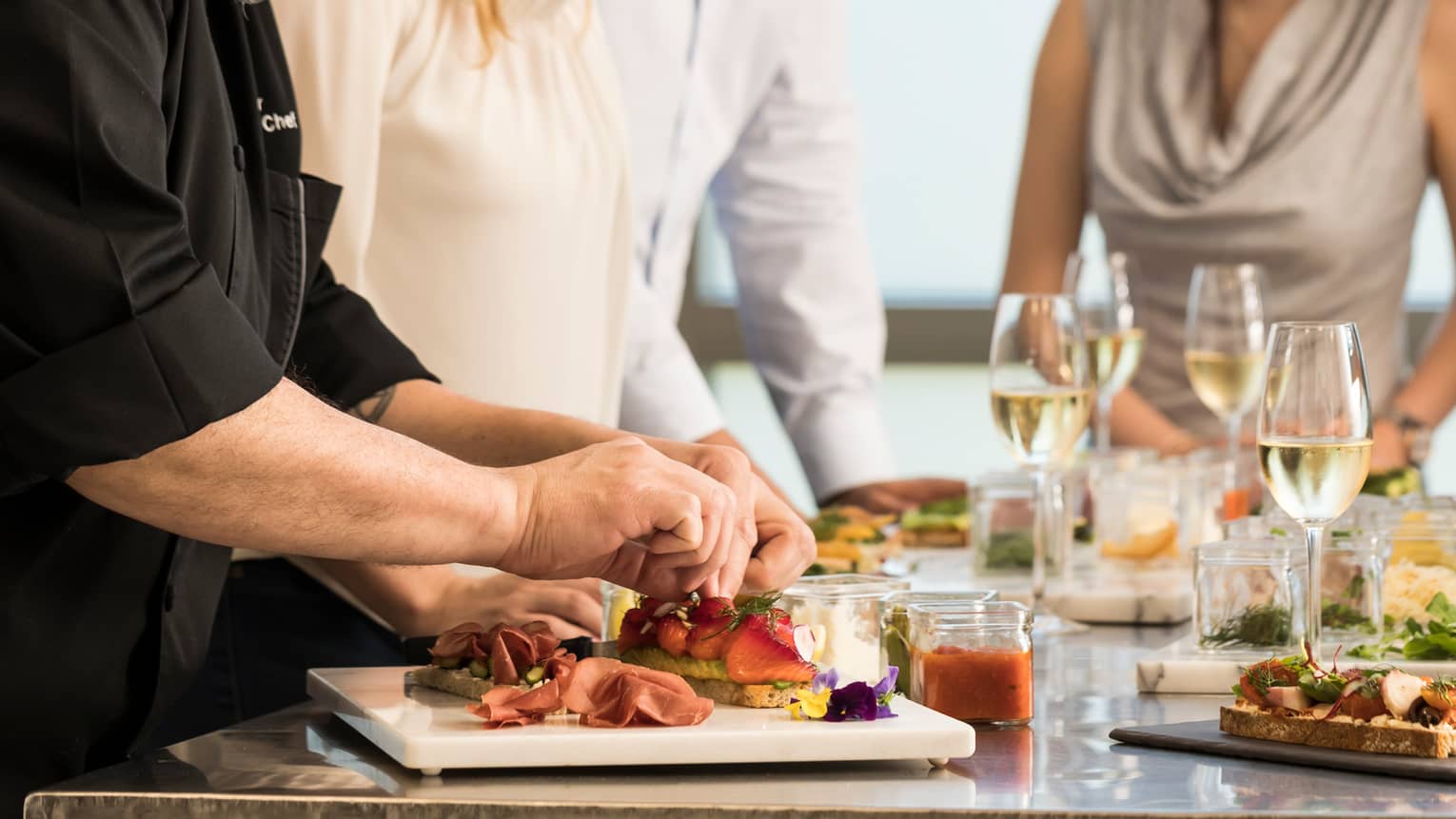 Chef assembles food at counter with smiling group of friends, white wine in glasses
