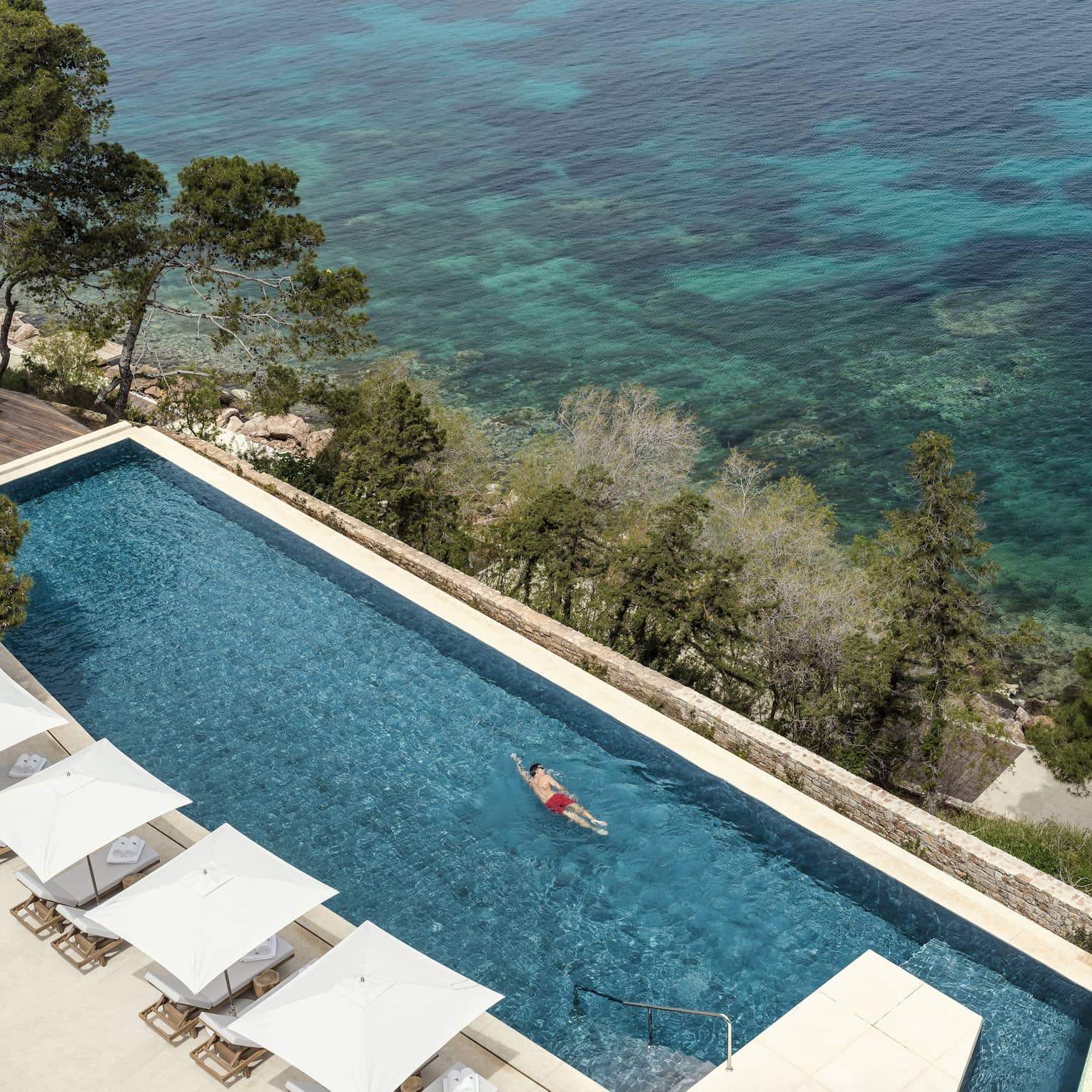 Man in red swim shorts swimming in infinity pool overlooking ocean and white cabanas 