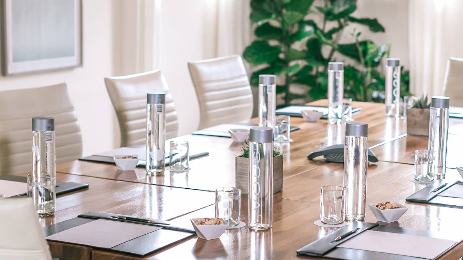 A rectangular boardroom table set with clear glass water bottles and black folders in a naturally-lit room