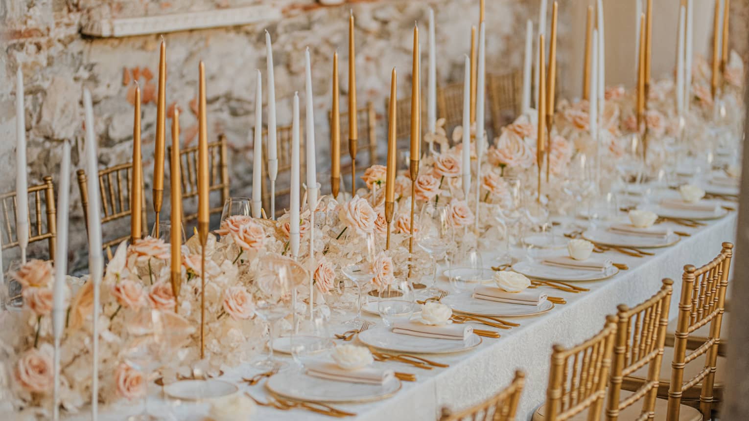 A long table in an indoor space, set for a celebration: white tablecloth, plates, napkins, utensils, white and peach floral arrangements all along the table centre, and white and peach taper candles in candlesticks