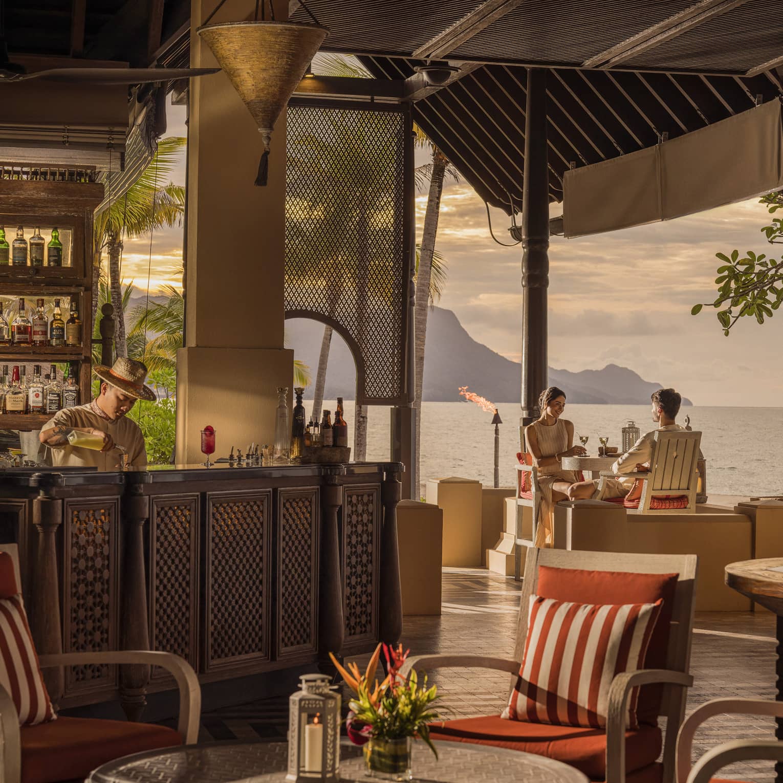 Two guests sips cocktails at open-air bar with ocean views while bartender makes drink on nearby bartop