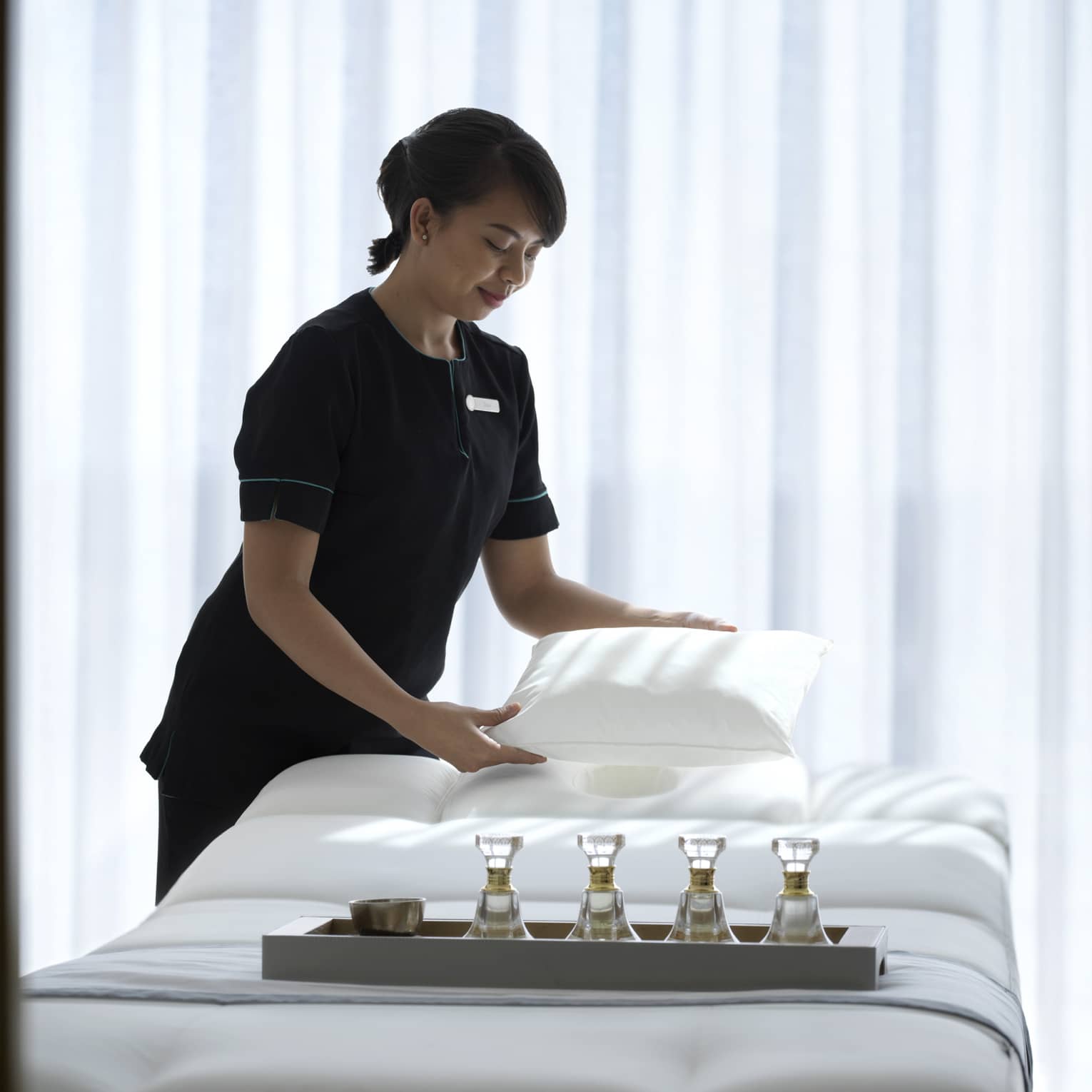 Spa attendant wearing a black uniform fluffs a while pillow on a treatment table covered with all-white linens and a tray filled with four small glass bottles