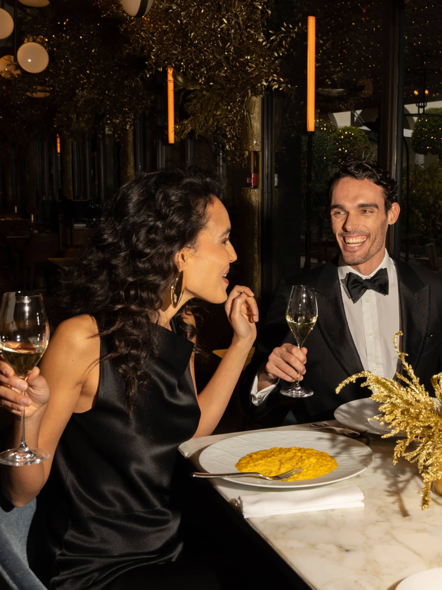 Three friends laugh together as they eat and hold champagne glasses at a table adorned with golden centrepieces.