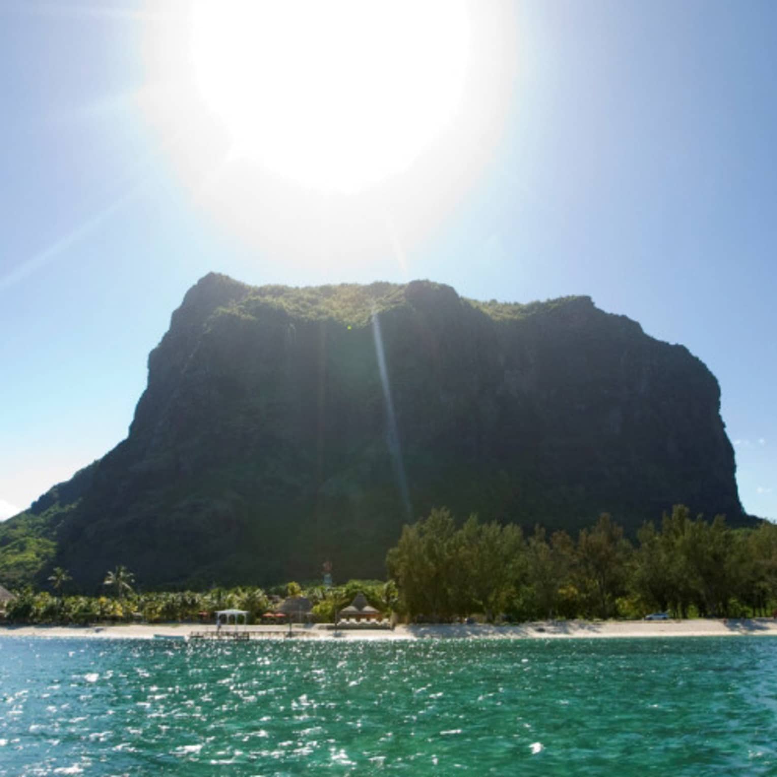 Long view of a forested monolith mountain sitting atop crystal waters. Sun beams down from above in a clear blue sky.