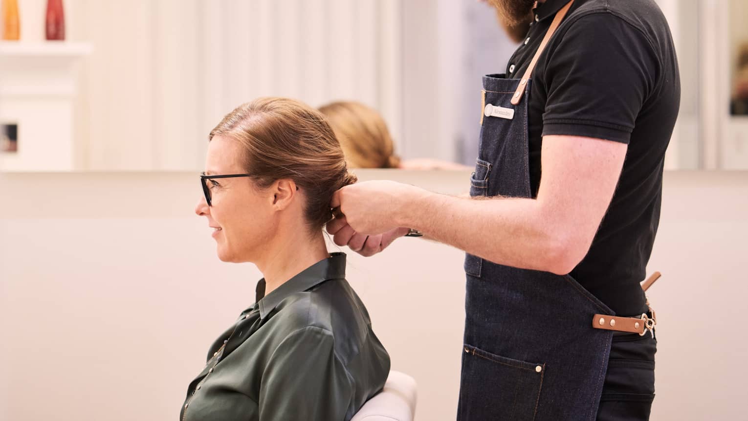 Woman sits in salon chair as hair stylist with long beard holds her hair